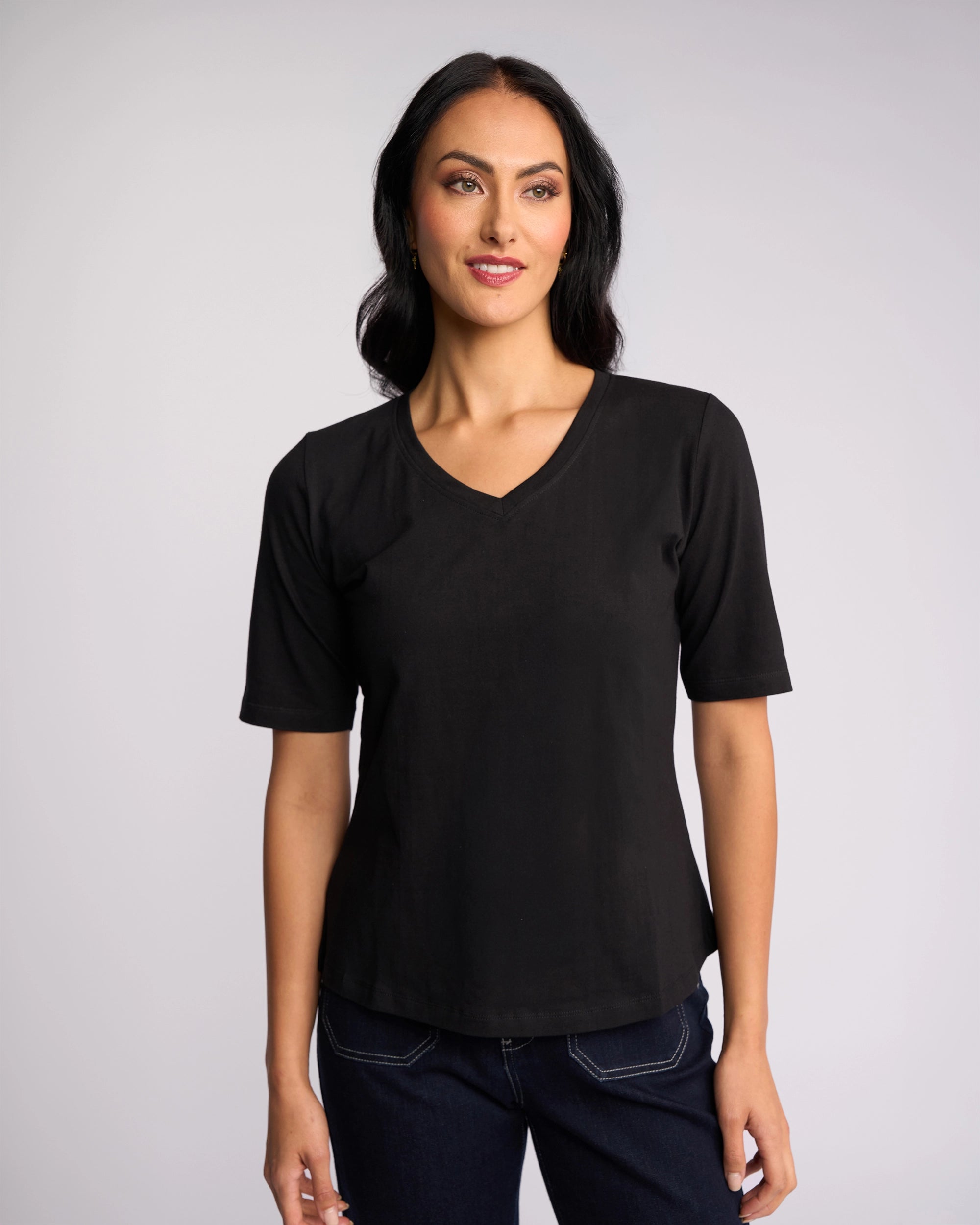 A woman with long dark hair, wearing a Vassalli Base Tee - Black and dark jeans, stands against a plain light background, smiling slightly.