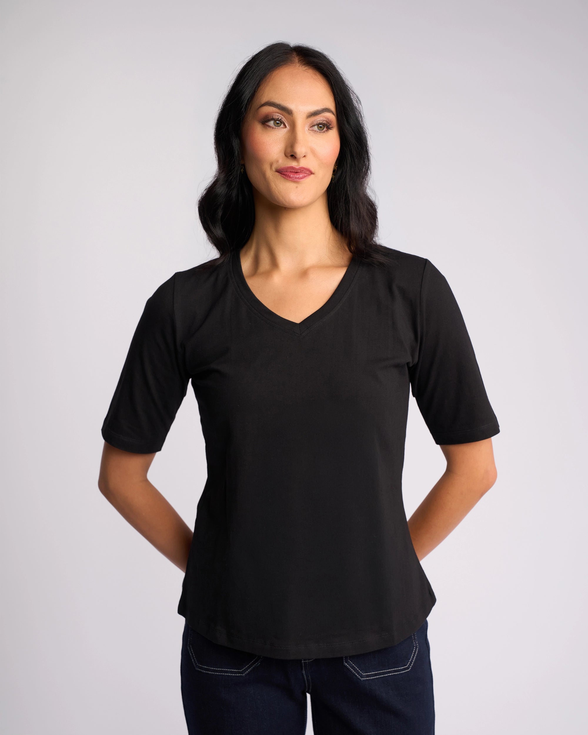 A woman with long dark hair wears the Vassalli Base Tee - Black with dark jeans, standing against a plain light background, hands behind her back, looking slightly to the side and smiling.