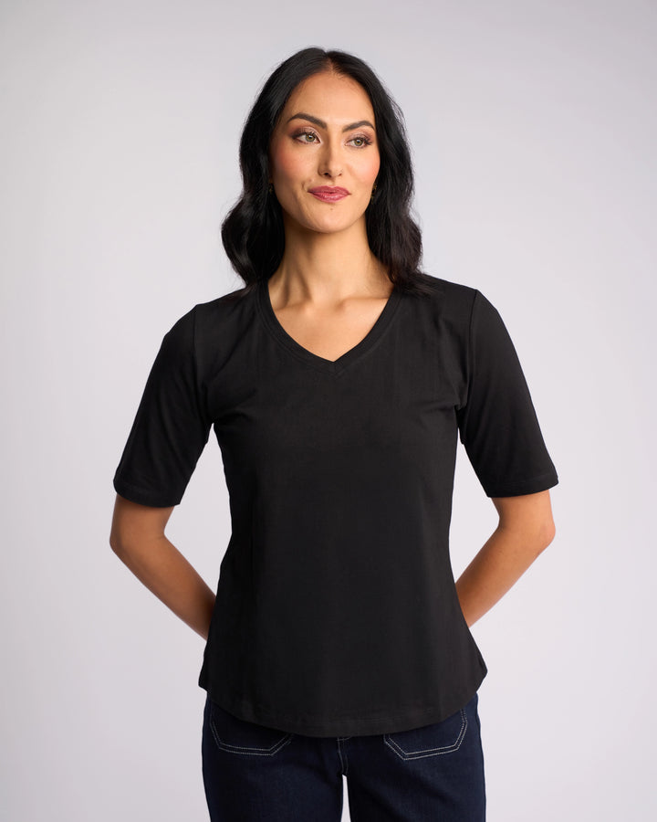 A woman with long dark hair wears the Vassalli Base Tee - Black with dark jeans, standing against a plain light background, hands behind her back, looking slightly to the side and smiling.
