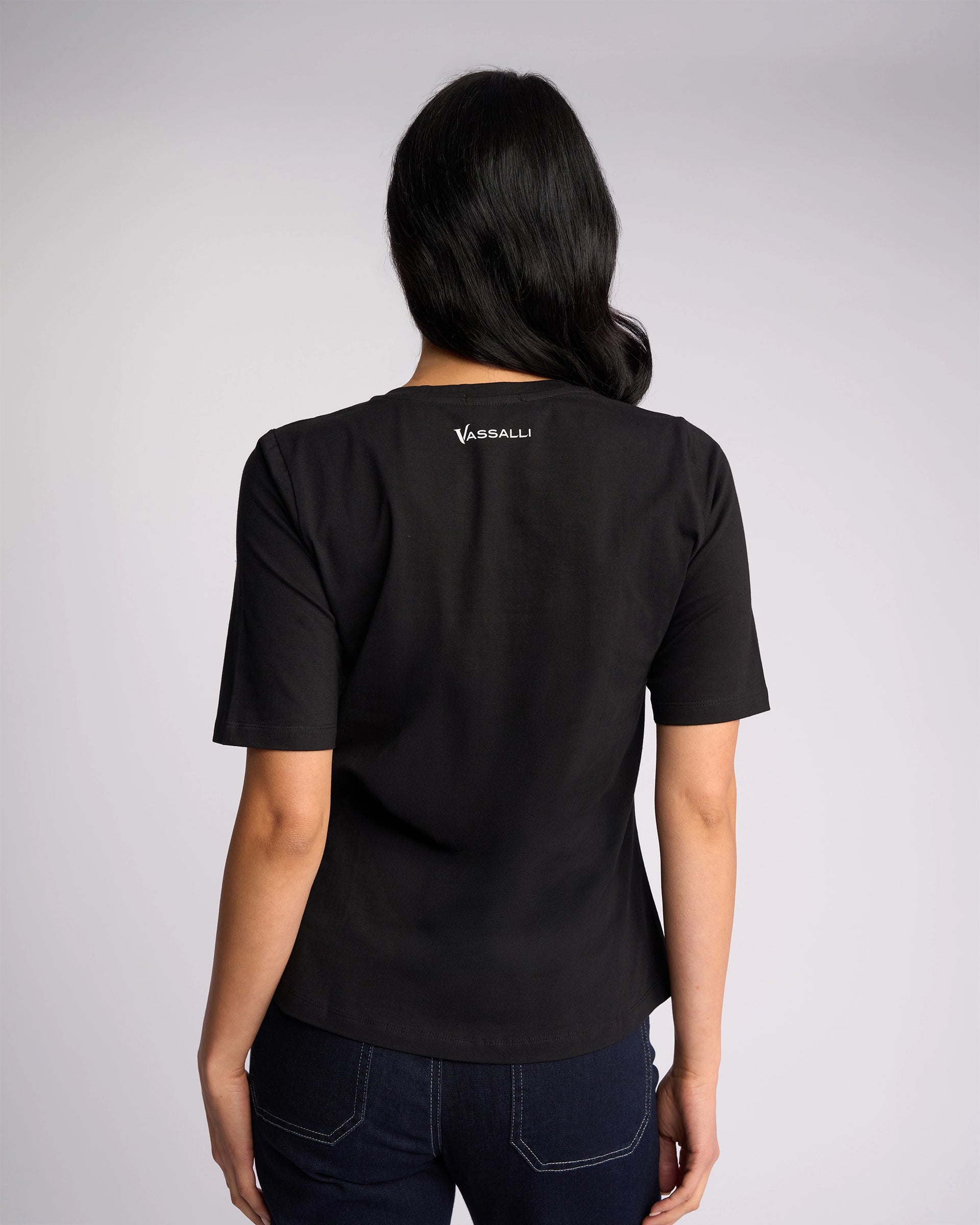 A woman stands with her back to the camera, wearing a Vassalli Base Tee - Black and dark blue jeans against a plain light background.