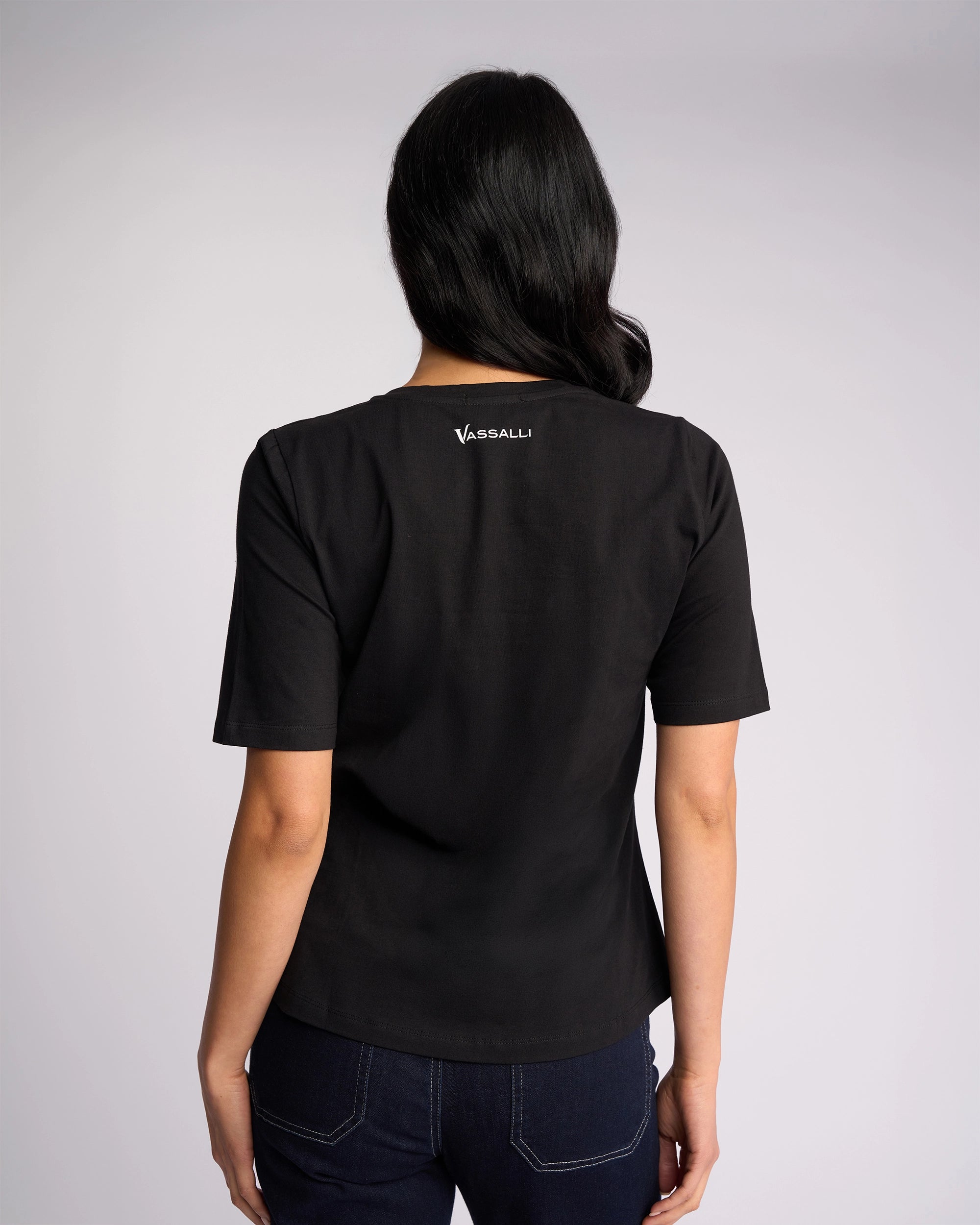 A woman stands with her back to the camera, wearing a Vassalli Base Tee - Black and dark blue jeans against a plain light background.