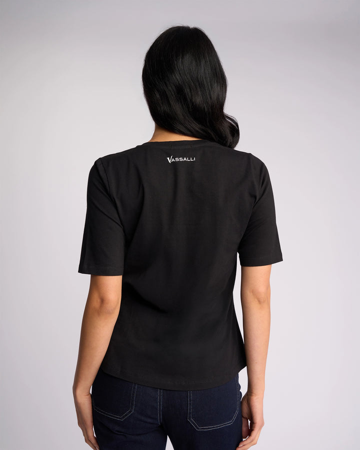 A woman stands with her back to the camera, wearing a Vassalli Base Tee - Black and dark blue jeans against a plain light background.