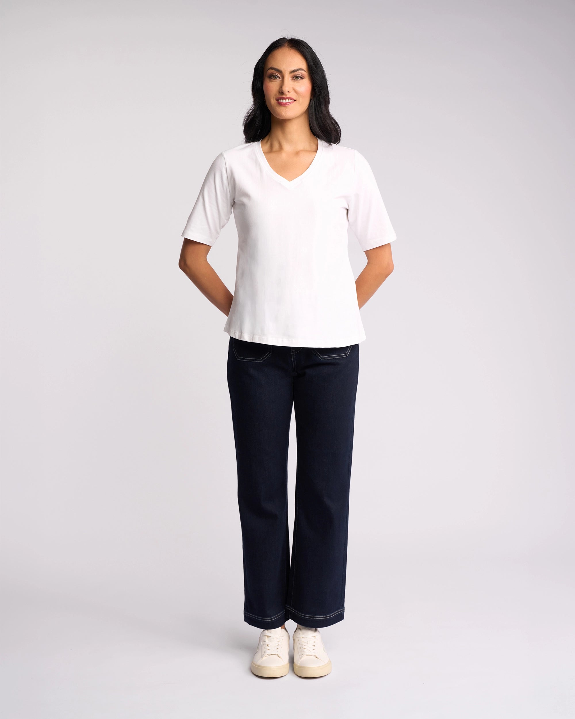 A woman faces forward with her hands behind her back, wearing the Vassalli Base Tee - White, dark straight-leg jeans, and white sneakers. The background is plain and light-colored.
