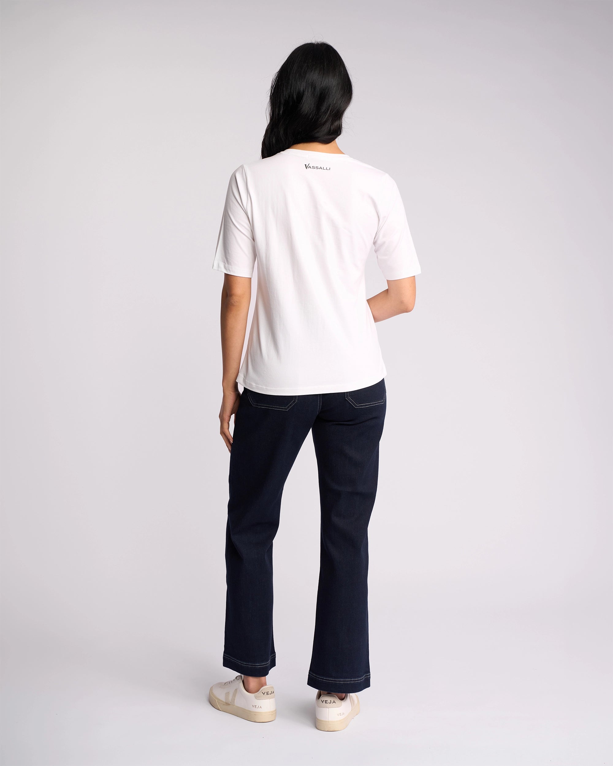 A person with long dark hair wears the Vassalli Base Tee in white, paired with dark jeans and white sneakers, standing with their back to a plain light background.