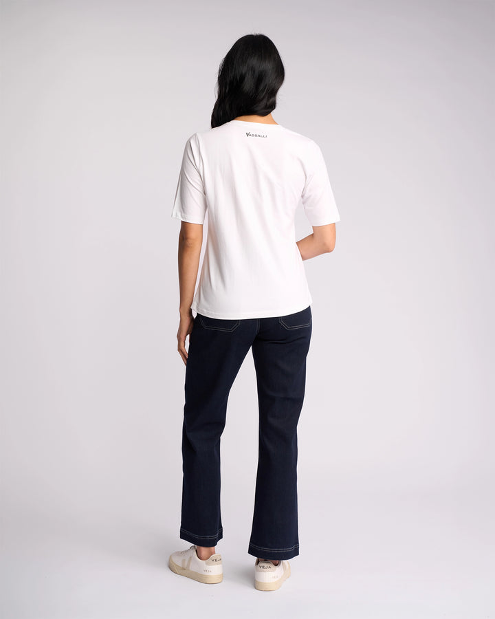 A person with long dark hair wears the Vassalli Base Tee in white, paired with dark jeans and white sneakers, standing with their back to a plain light background.
