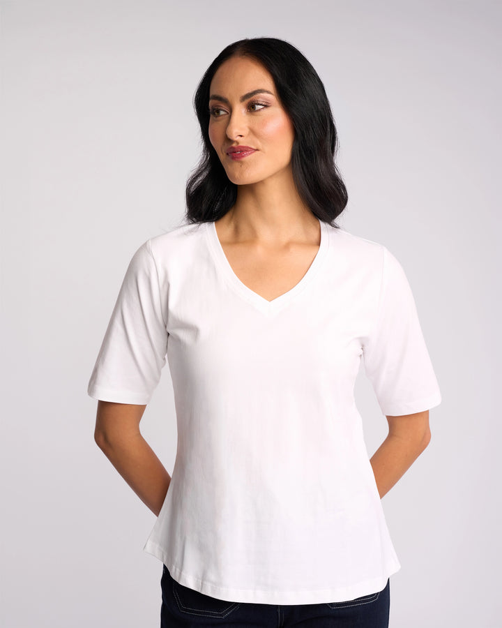 A woman with long dark hair stands against a plain light background, wearing the Vassalli Base Tee - White with a V-neckline and dark pants, looking slightly to her left with her hands behind her back.