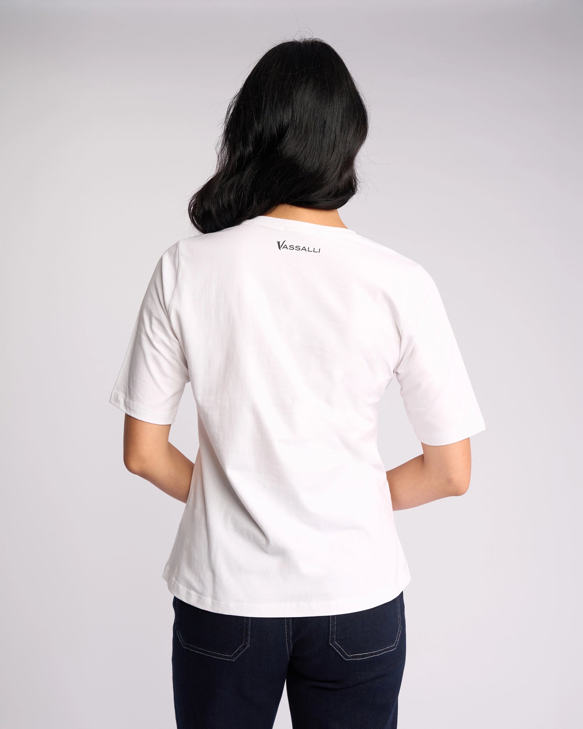 A person with long dark hair faces away, wearing the Vassalli Base Tee - White in 100% cotton with "Vassalli" on the upper back, paired with dark jeans against a plain light background.
