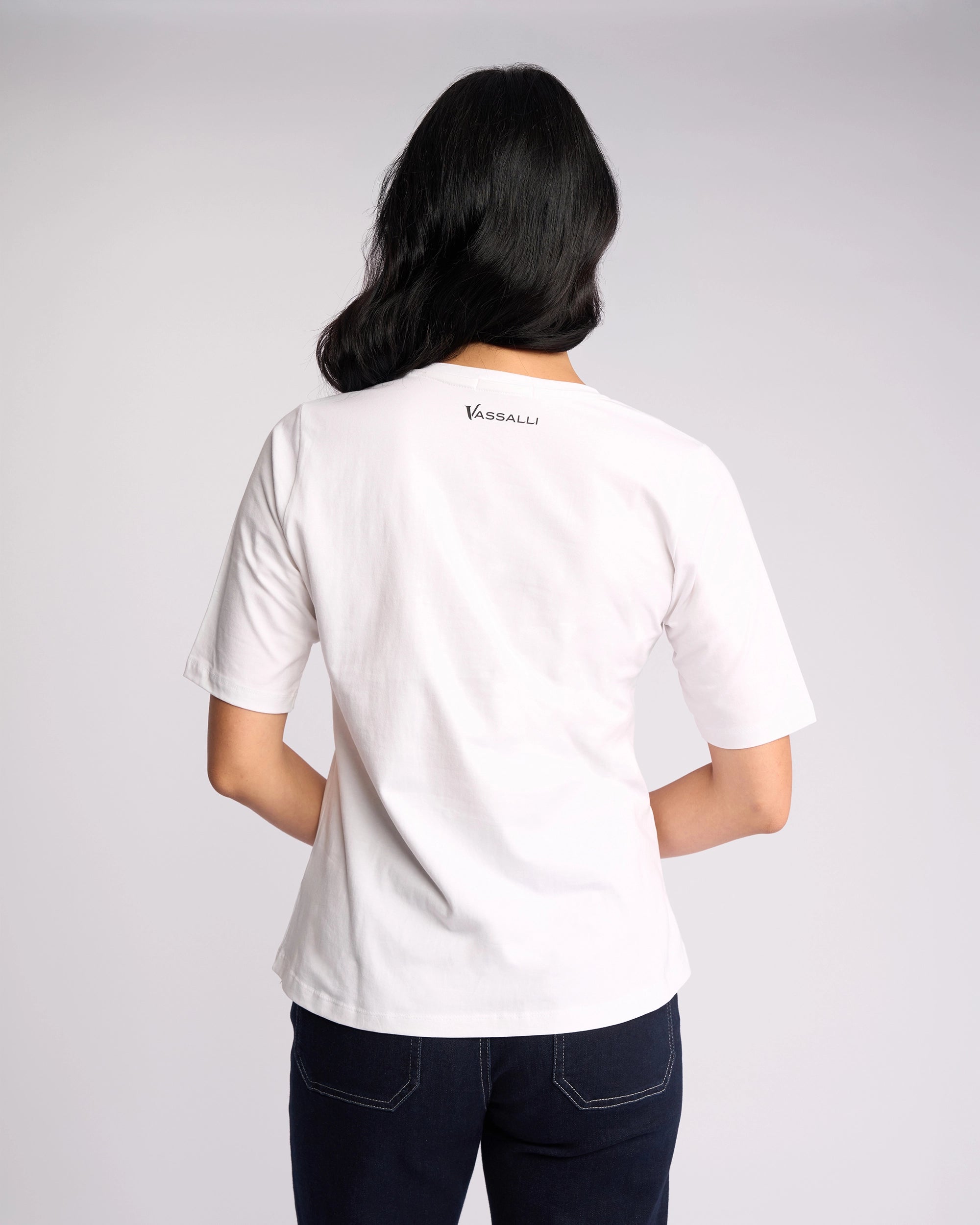 A person with medium-length dark hair is shown from the back, wearing a white Vassalli Base Tee featuring a V-neckline and "VASSALLI" on the upper back, paired with dark jeans against a light plain background.