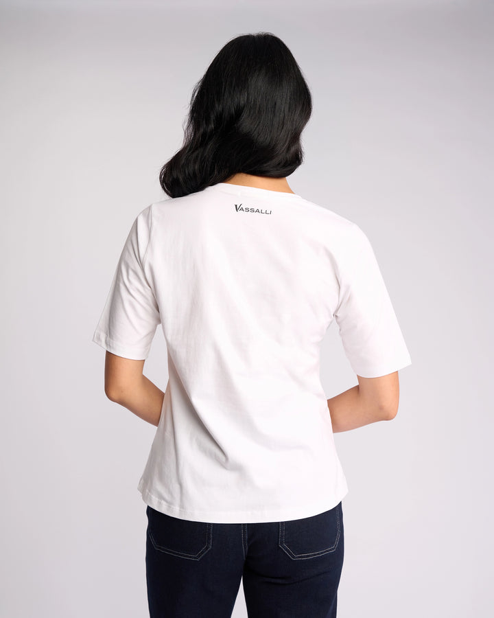 A person with medium-length dark hair is shown from the back, wearing a white Vassalli Base Tee featuring a V-neckline and "VASSALLI" on the upper back, paired with dark jeans against a light plain background.