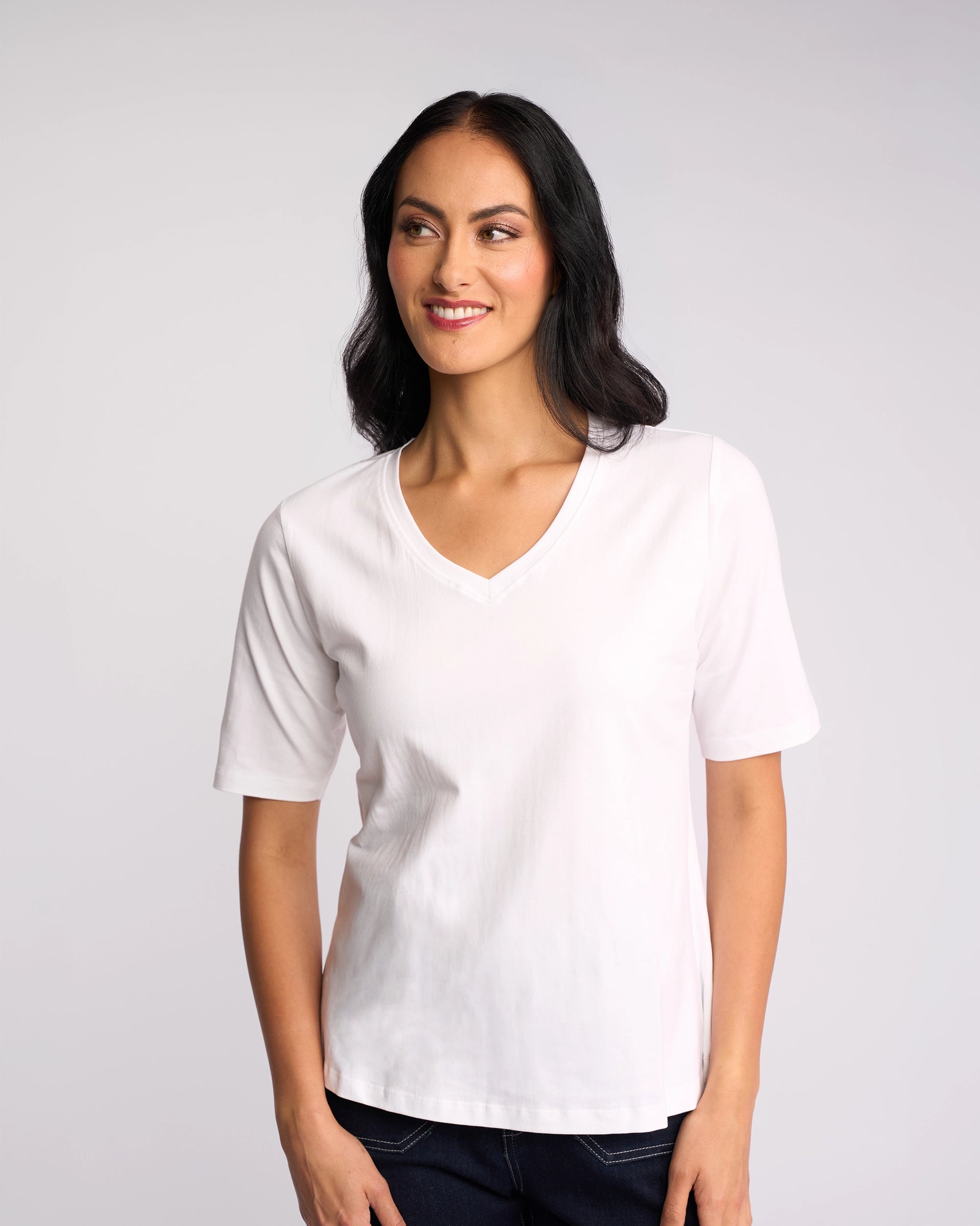 A woman with long dark hair, wearing the Vassalli Base Tee - White and dark pants, stands against a light gray background, smiling and glancing to the side.