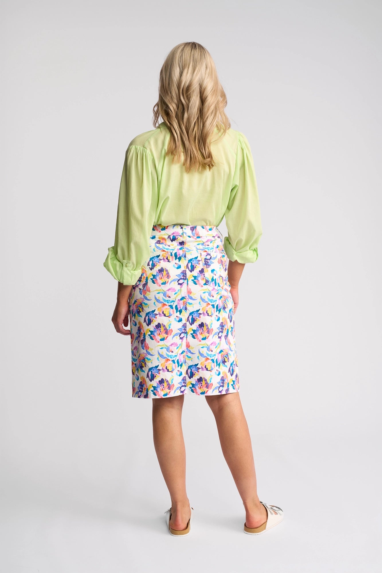 A person with wavy blonde hair faces away, wearing a light green long-sleeve blouse and the Vassalli Poppy Skirt - Demi, which features a colorful floral print. They stand against a plain white background, with white shoes visible.