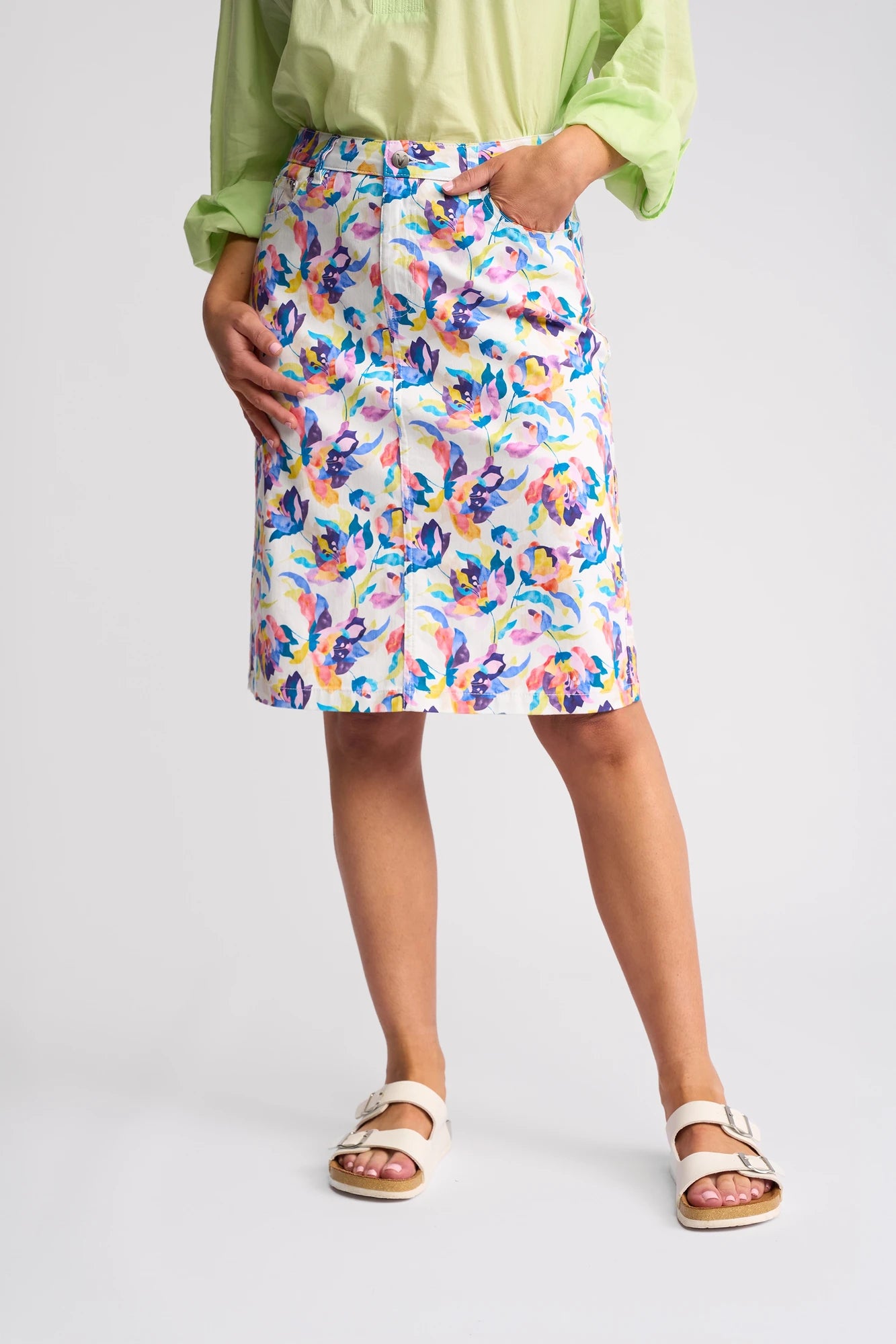 A person is wearing a light green blouse, the Vassalli Poppy Skirt - Demi featuring a colorful floral print in a knee-length summer style, and white sandals. They are standing with one hand in the skirt pocket against a plain background.