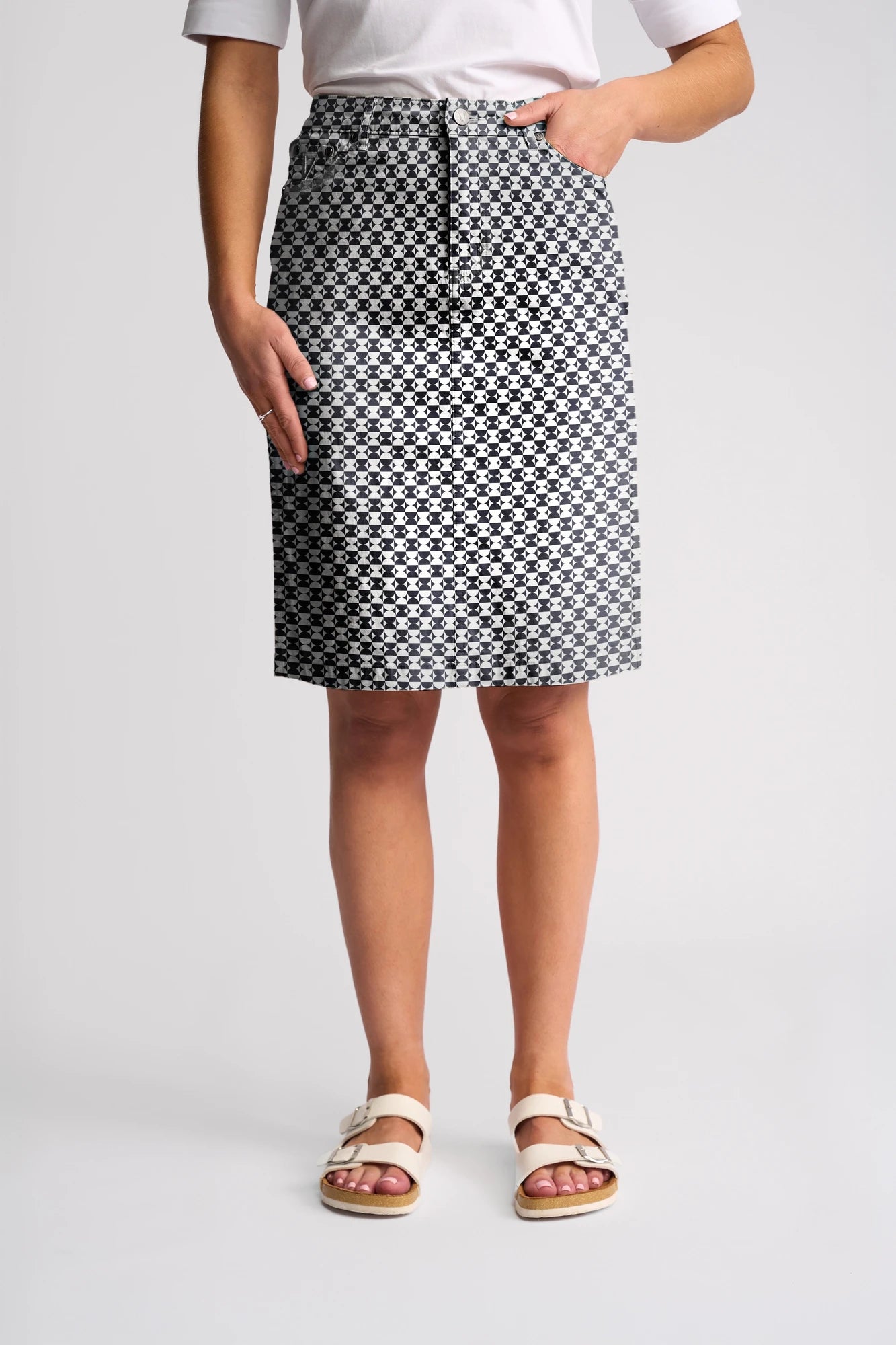 A person wearing a white top, the Vassalli Poppy Skirt - Dixie featuring a knee-length black and white geometric pattern, and white sandals stands with one hand in their pocket against a plain background.