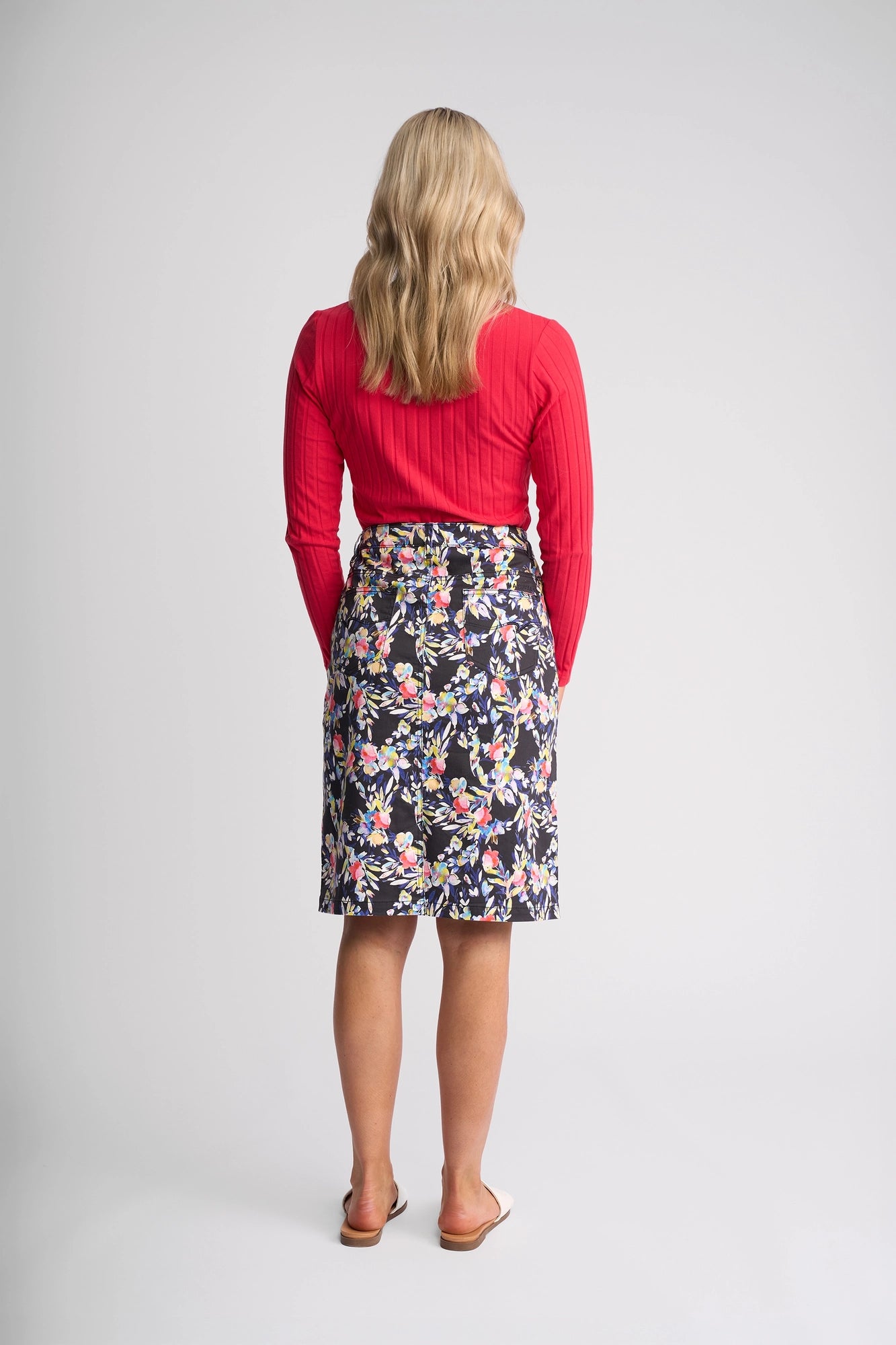 A woman with wavy blonde hair stands facing away, wearing a long-sleeve red top, the Vassalli Poppy Skirt - Lani—a knee-length floral print skirt with a black background—and white sandals. The background is plain and light gray.