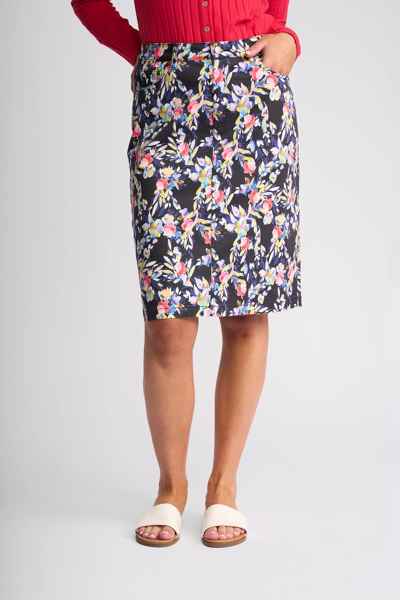A person wearing the Vassalli Poppy Skirt - Lani, paired with a red top and white open-toe sandals, stands against a plain light background, with only the lower half of their body visible.