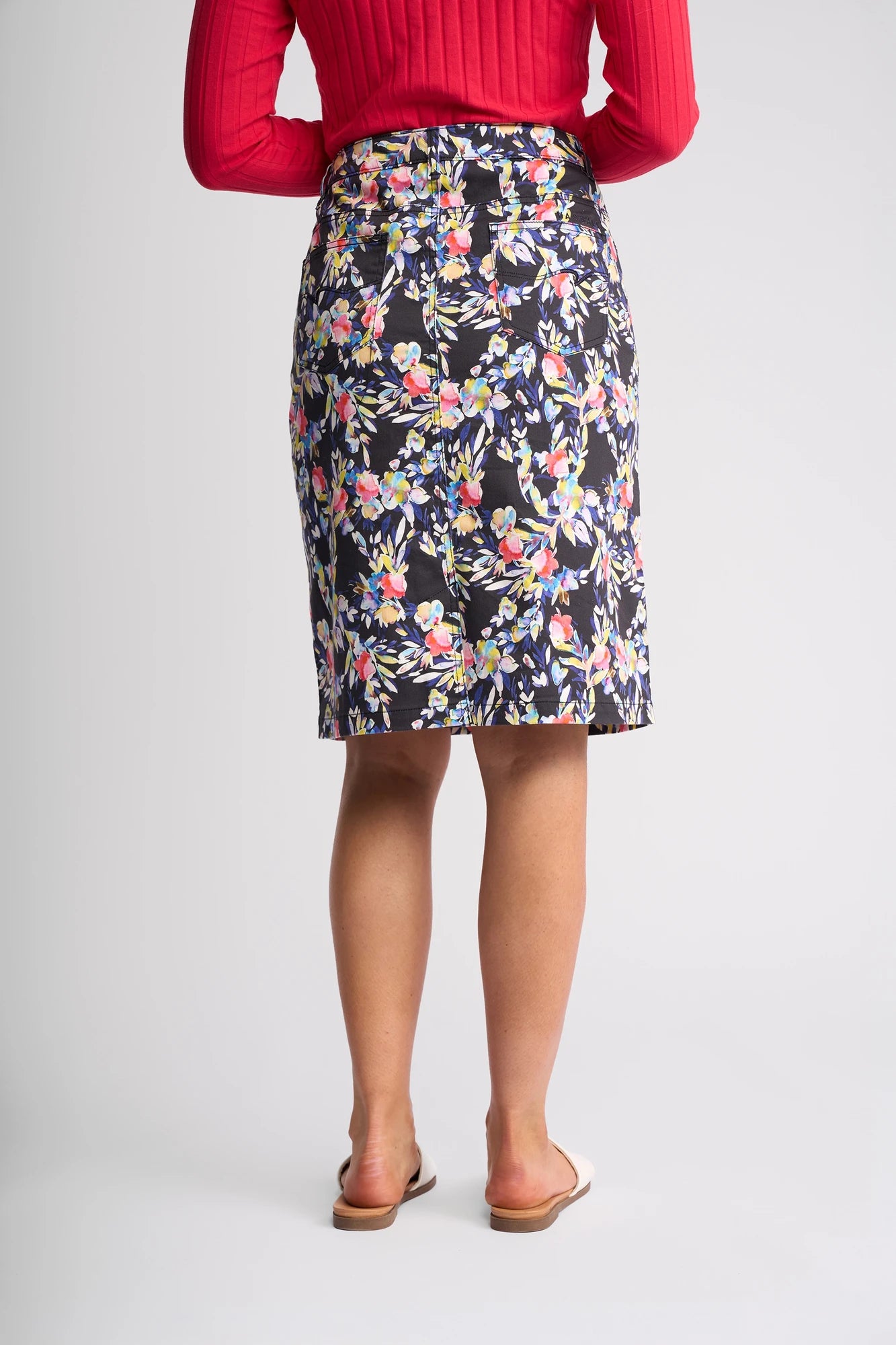 A person wearing a red long-sleeve top, the Vassalli Poppy Skirt - Lani (a knee-length dark floral print skirt), and tan sandals is standing facing away from the camera against a plain light background.