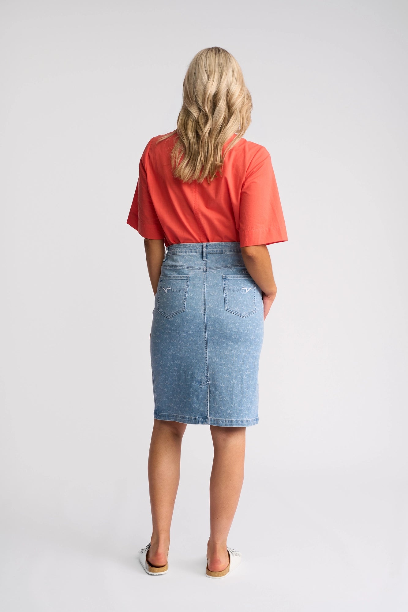 A woman with wavy blonde hair, wearing a red short-sleeve top and the Vassalli Poppy Skirt - Meadow Denim, stands facing away from the camera against a plain light background.
