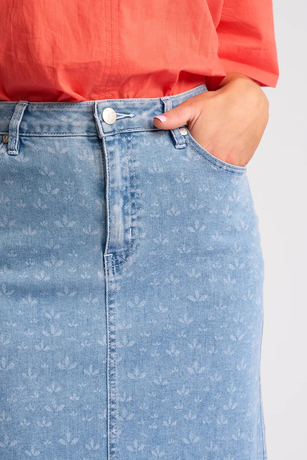 A person wearing a coral shirt and the Vassalli Poppy Skirt - Meadow Denim, with one hand in the front pocket; image cropped to show the waist and part of the torso.