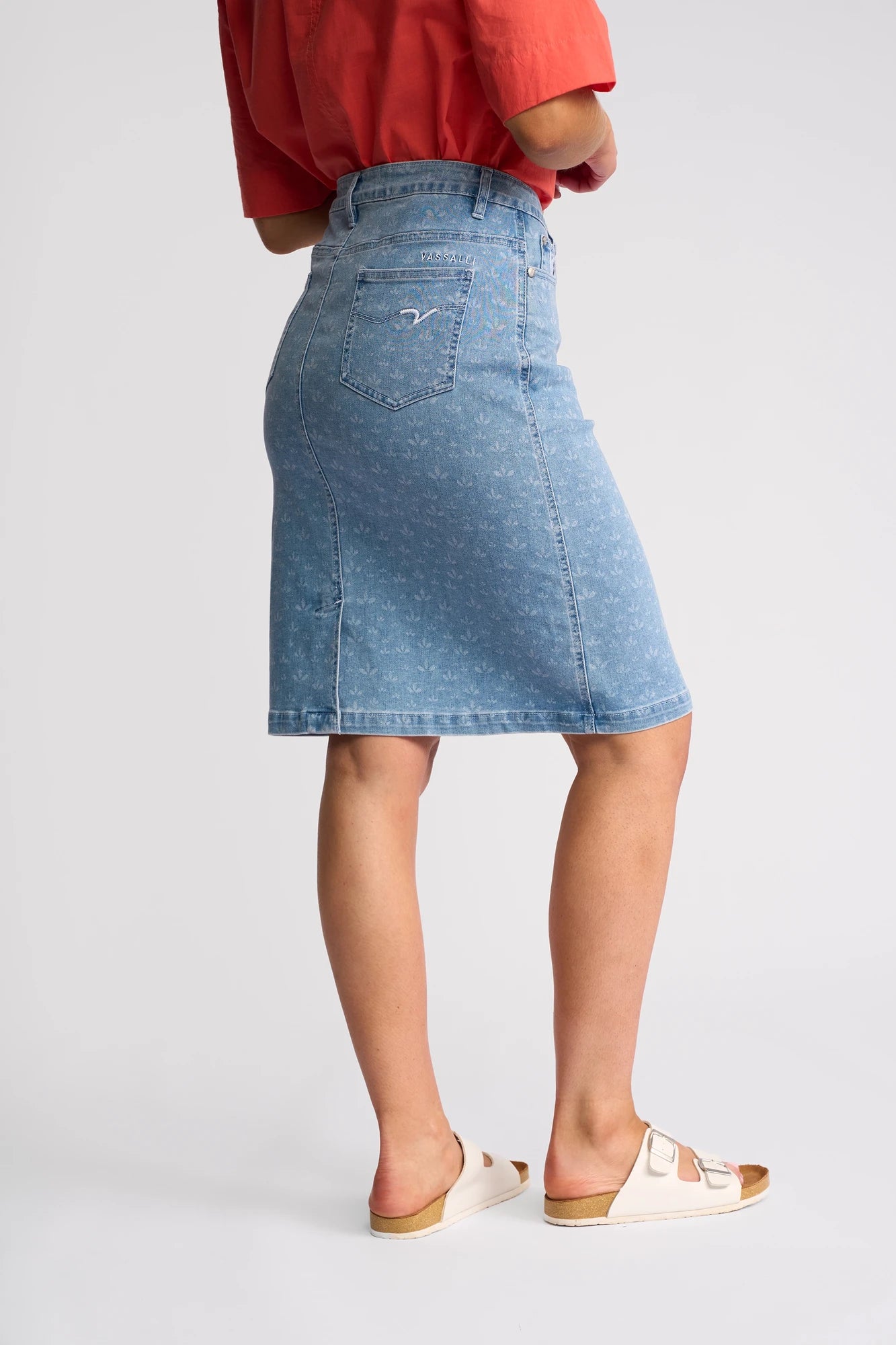 A person wearing a red shirt, white sandals, and the Vassalli Poppy Skirt - Meadow Denim—a blue stretch denim, knee-length pencil skirt with a back pocket—stands against a plain light background.
