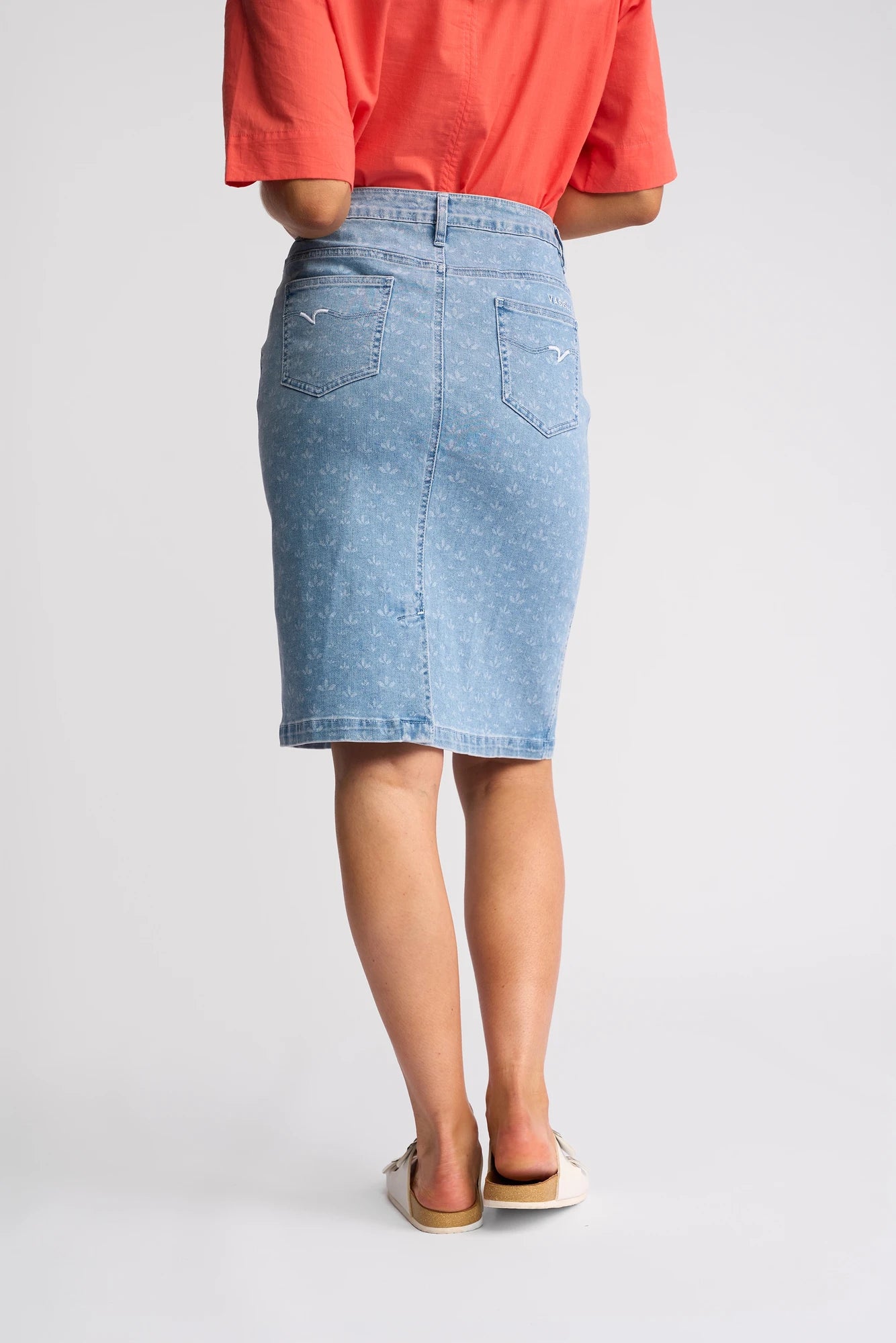 A person wearing the Vassalli Poppy Skirt - Meadow Denim, paired with a coral short-sleeve shirt and white slip-on shoes, stands facing away from the camera against a plain background.