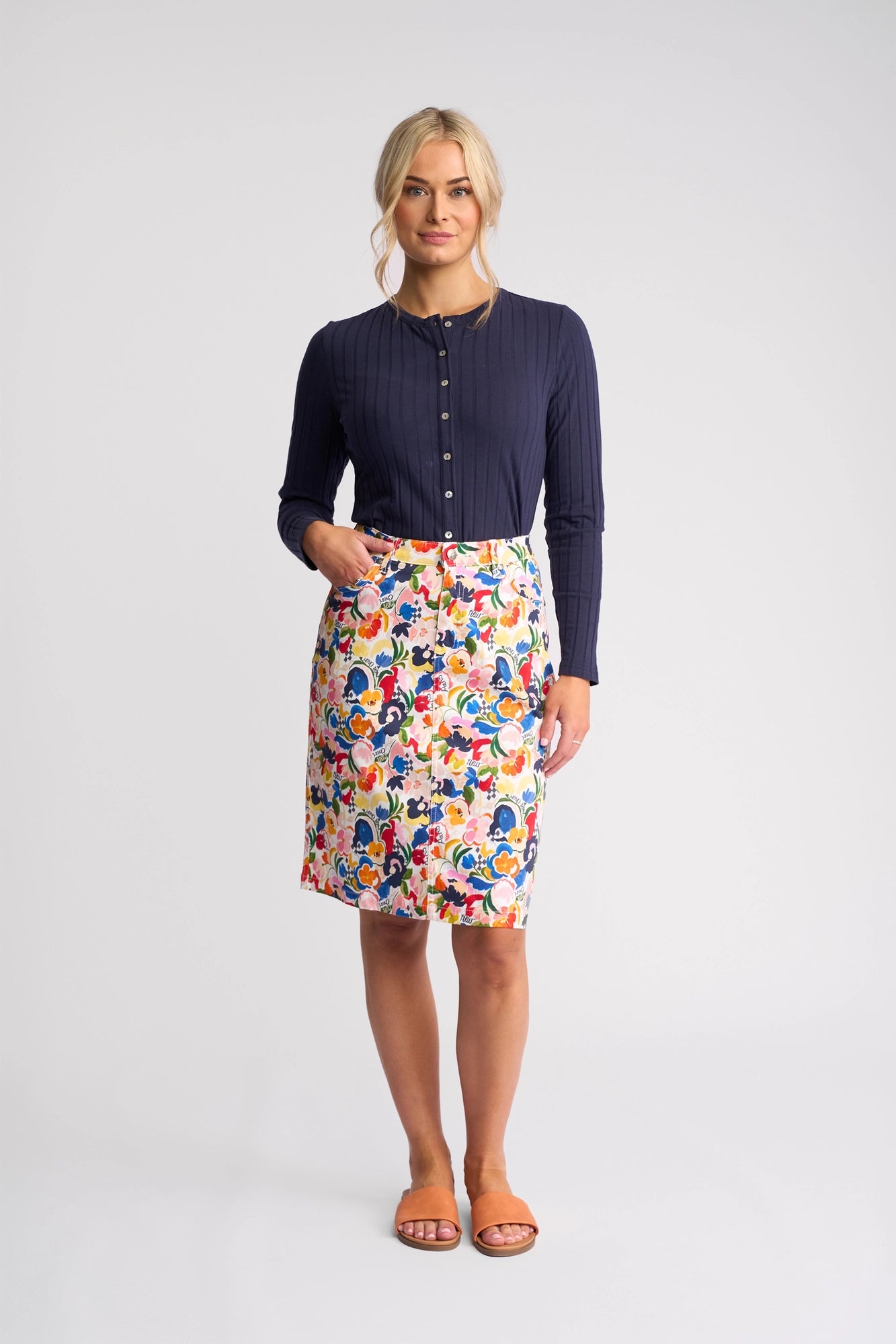 A woman with light hair tied back stands against a plain background, wearing the Vassalli Poppy Skirt - Mon Cheri, paired with a navy long-sleeve buttoned top and tan sandals. She is looking forward and smiling slightly.