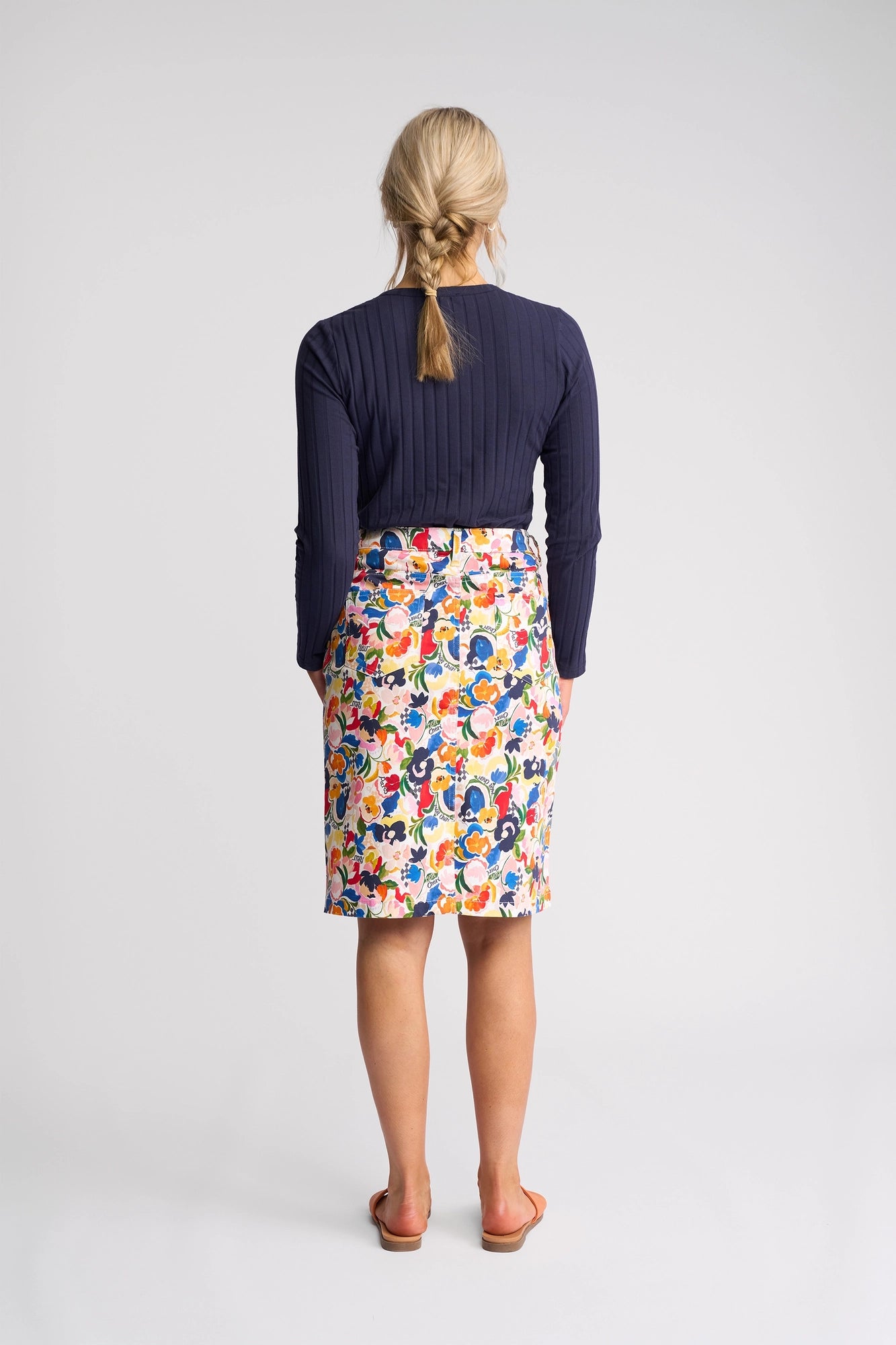 A woman with blonde hair in a braid, dressed in a navy long-sleeve top, the Vassalli Poppy Skirt - Mon Cheri, and sandals, stands facing away from the camera against a plain white background.