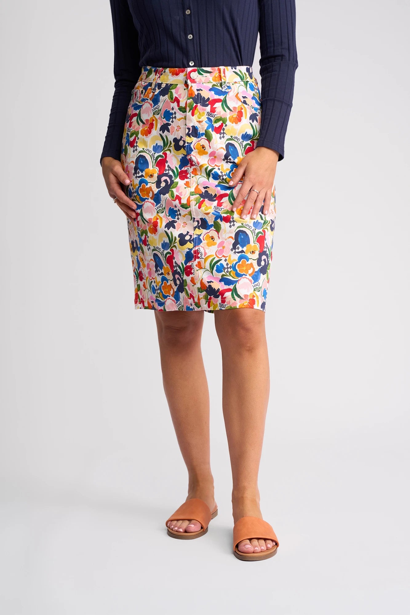 A person wearing a navy long-sleeve top, Vassalli’s Poppy Skirt - Mon Cheri featuring a vibrant floral print that falls to the knee, and orange slide sandals stands against a plain background.