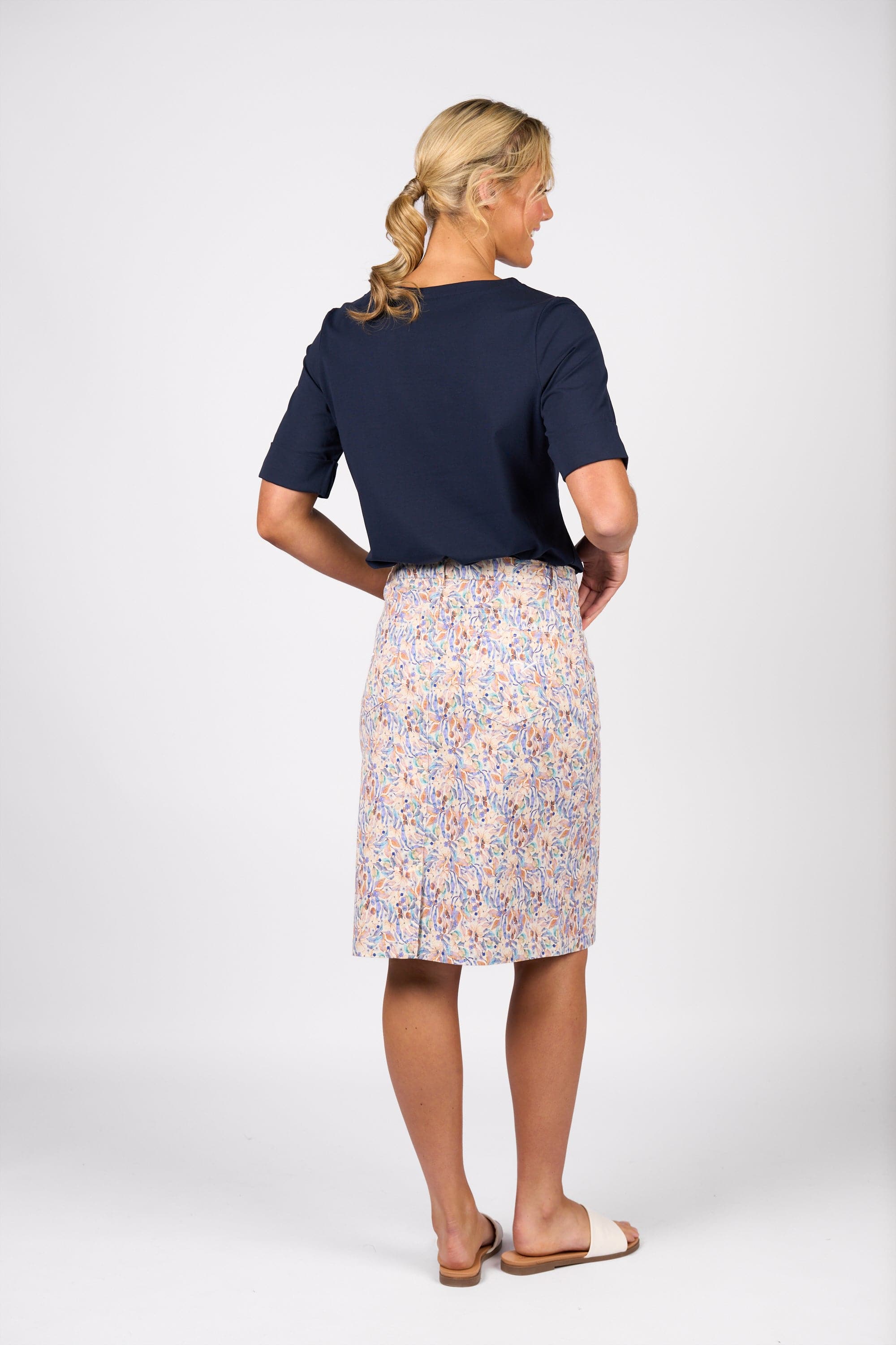 A woman with blonde hair in a ponytail stands facing away, wearing a navy top, the Vassalli Poppy Skirt - Amalfi Denim, and white sandals against a plain white background.