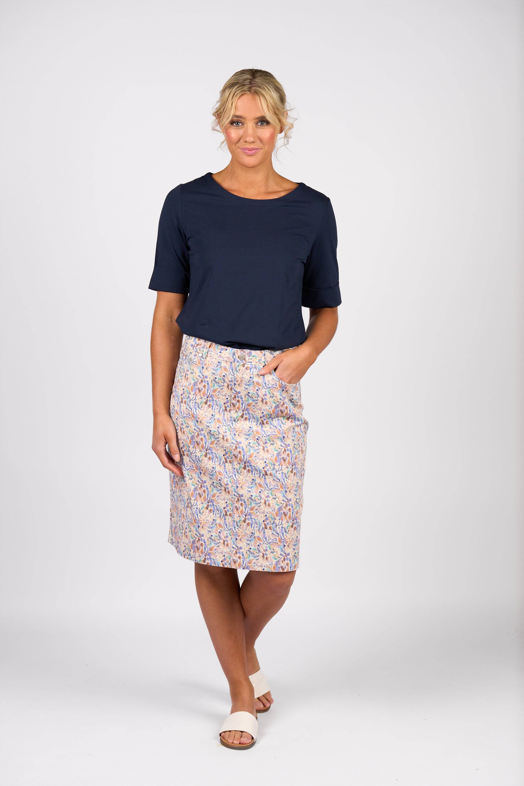 A woman with blonde hair in an updo poses against a white background, wearing the Vassalli Poppy Skirt - Amalfi Denim with a navy short-sleeve top and white slide sandals, her hand resting in her pocket.