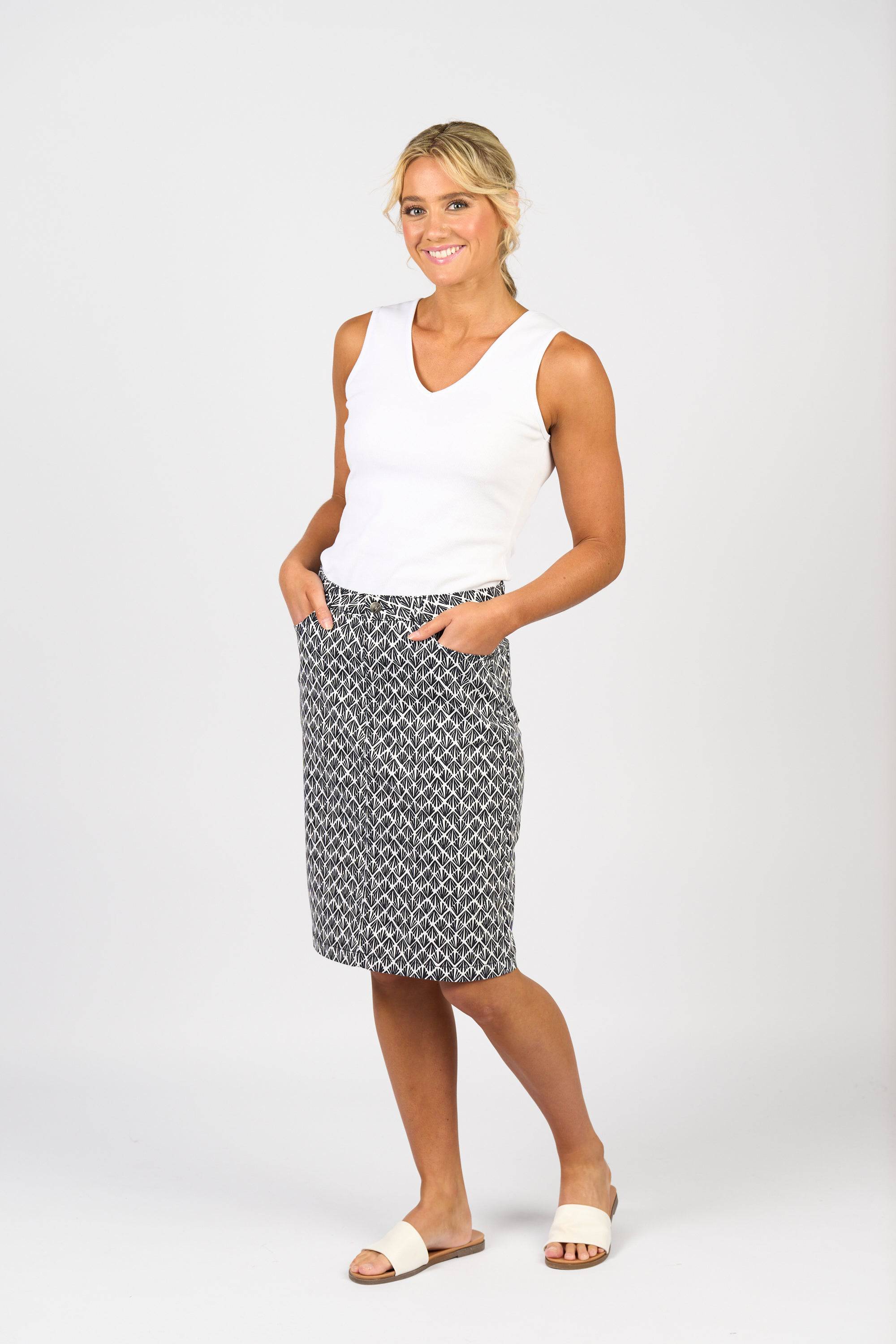 A woman smiles with her hands in the pockets of her Vassalli Poppy Lightweight Skirt - Fan, paired with a white sleeveless top and white slide sandals, creating an ideal everyday outfit against a plain light background.