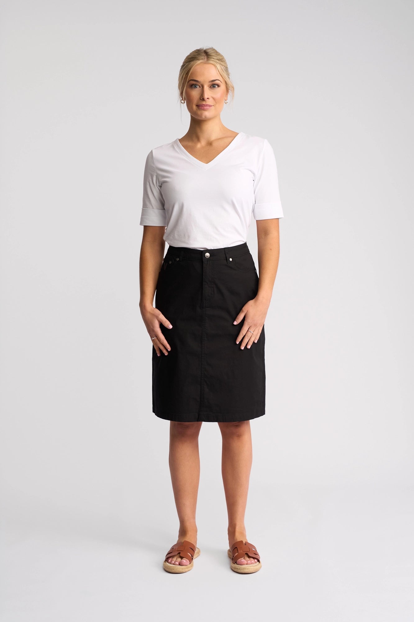 A woman stands facing forward against a plain light background, wearing the Vassalli Poppy Poplin Skirt in black, paired with a white V-neck shirt and brown sandals. She has a neutral expression.