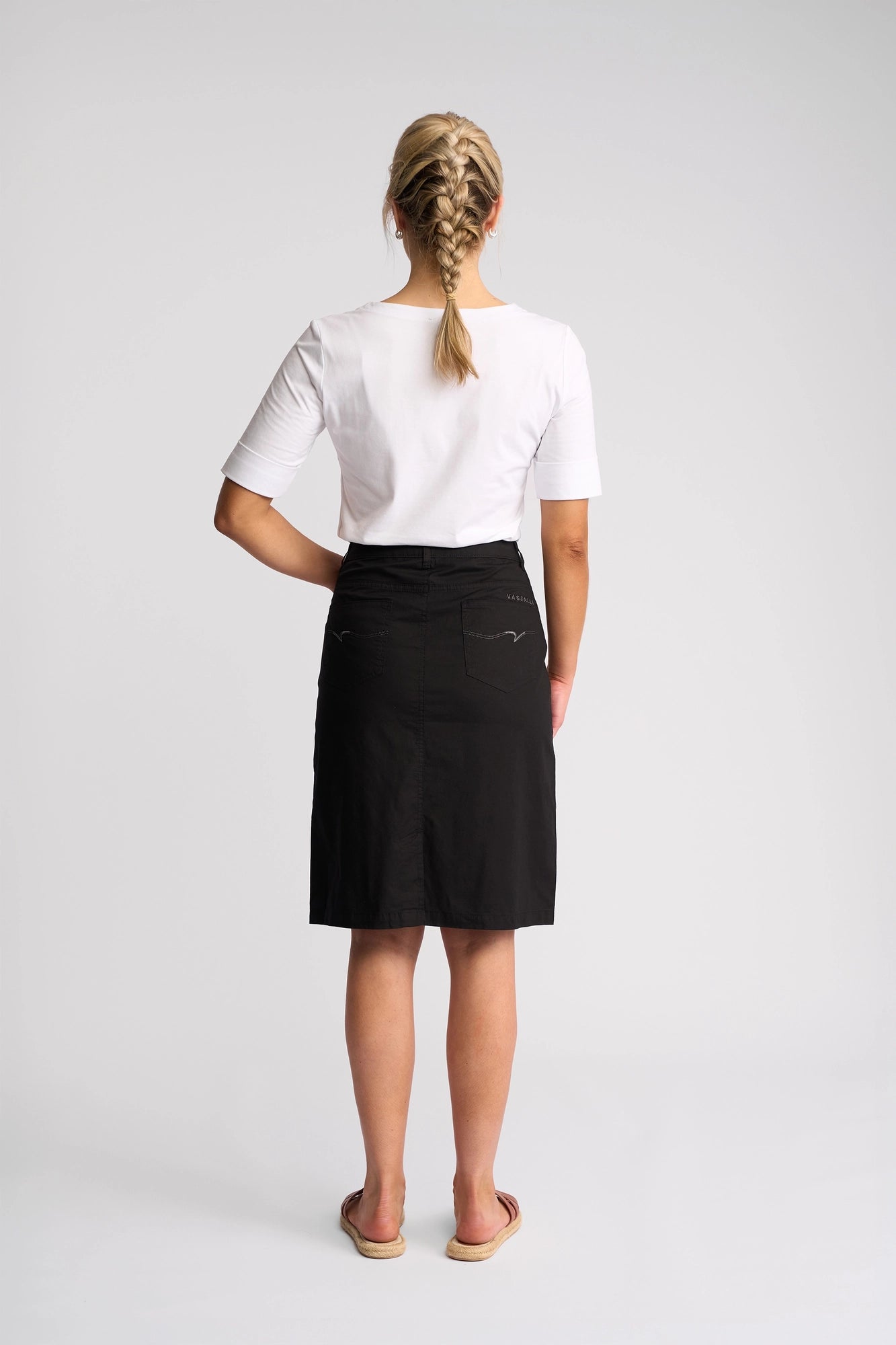 A woman with blonde braided hair stands facing away, wearing a white short-sleeve top, the Vassalli Poppy Poplin Skirt in black at knee length, and sandals, against a plain light background.