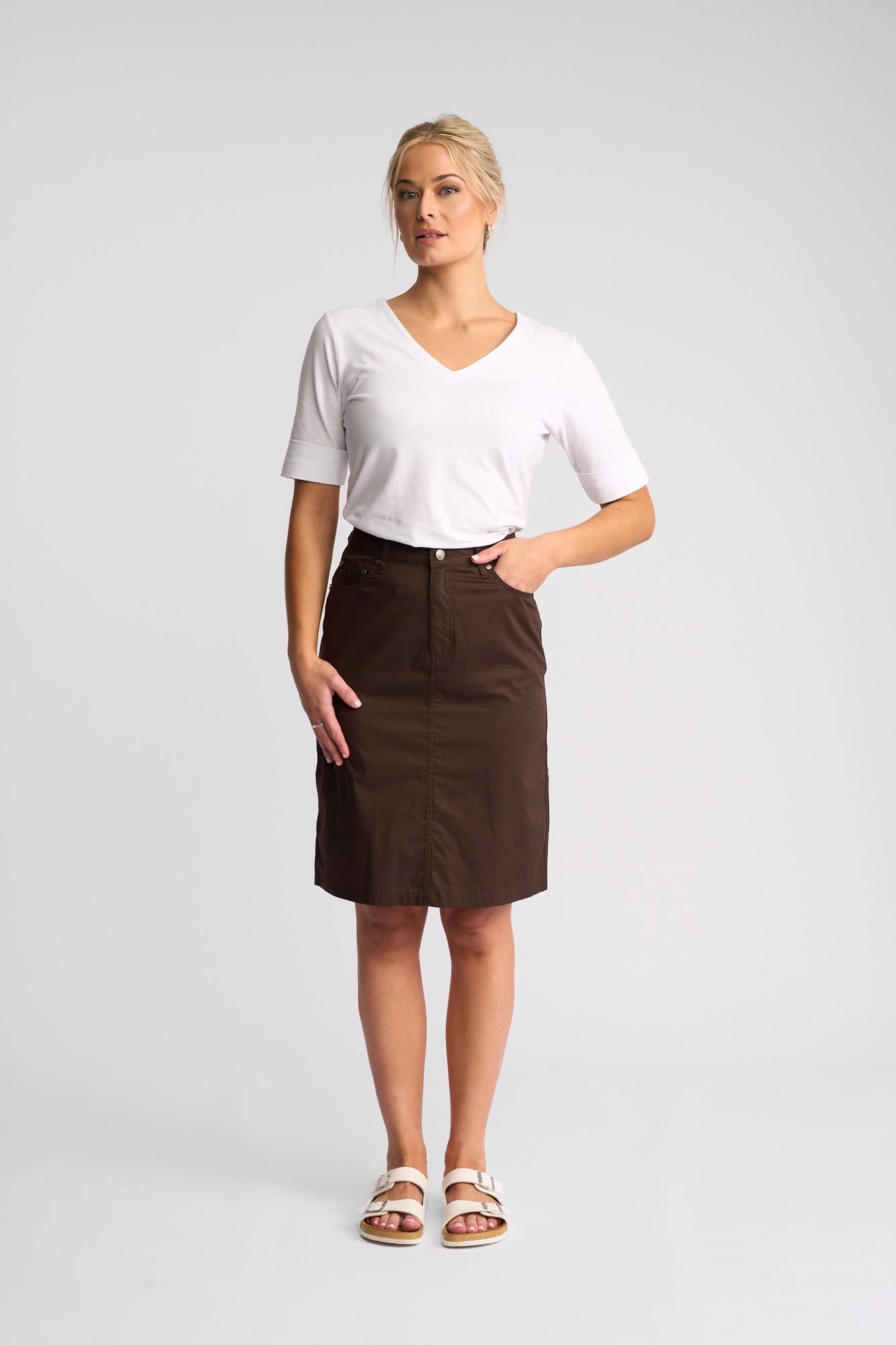 A person stands in a white studio wearing a crisp white V-neck T-shirt, the Vassalli Poppy Poplin Skirt in Chocolate, and white sandals. Their blonde hair is tied back, and they look slightly to the side with one hand on their hip.