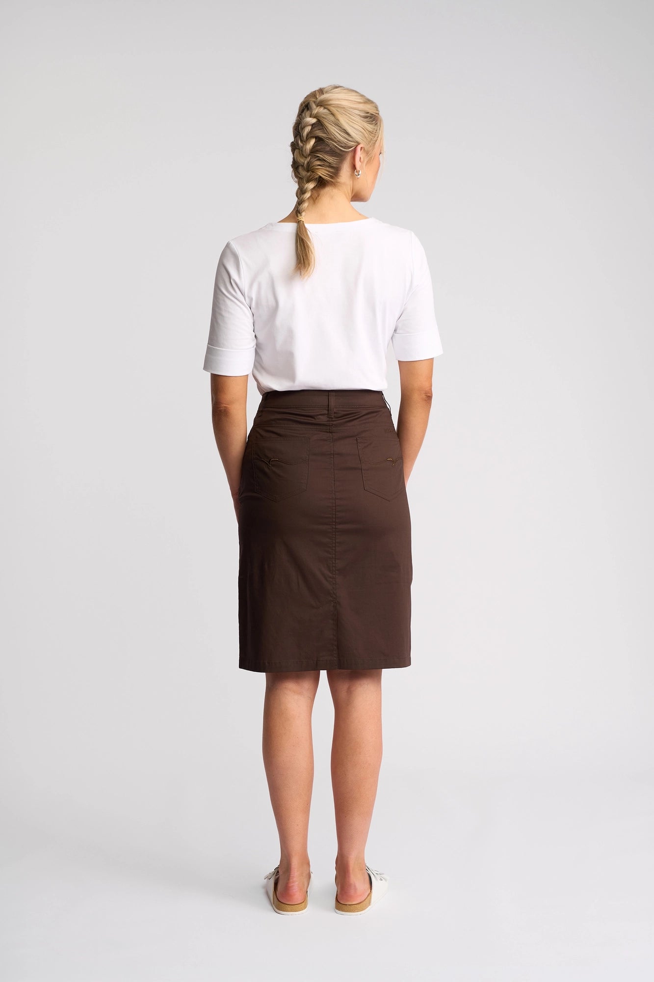 A woman with blonde hair in two braids stands facing away, wearing a white short-sleeve shirt, the Vassalli Poppy Poplin Skirt in Chocolate, and white shoes against a plain light background.