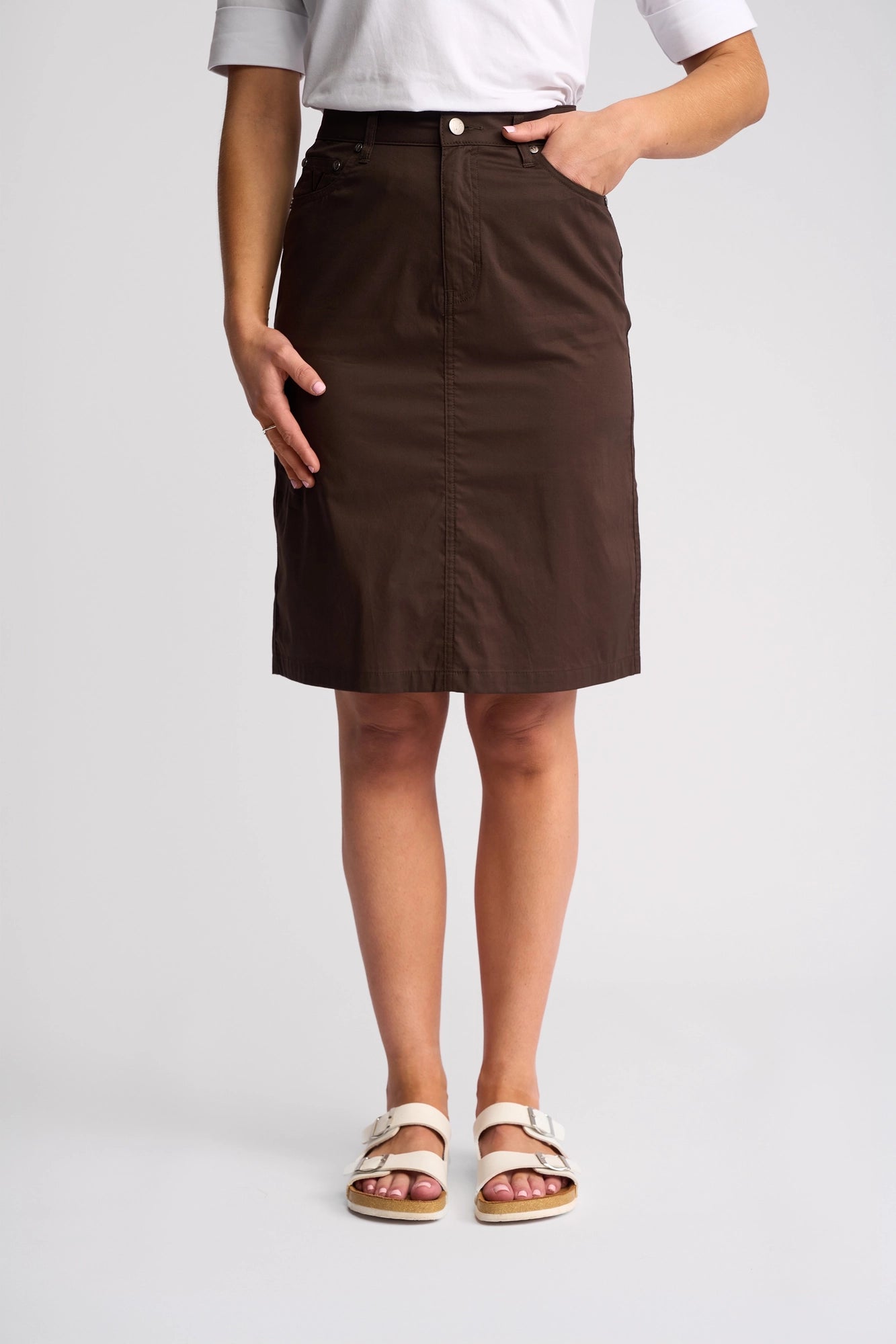 A person wearing a white top, the Vassalli Poppy Poplin Skirt in Chocolate, and white sandals stands with one hand in their skirt pocket against a plain background.