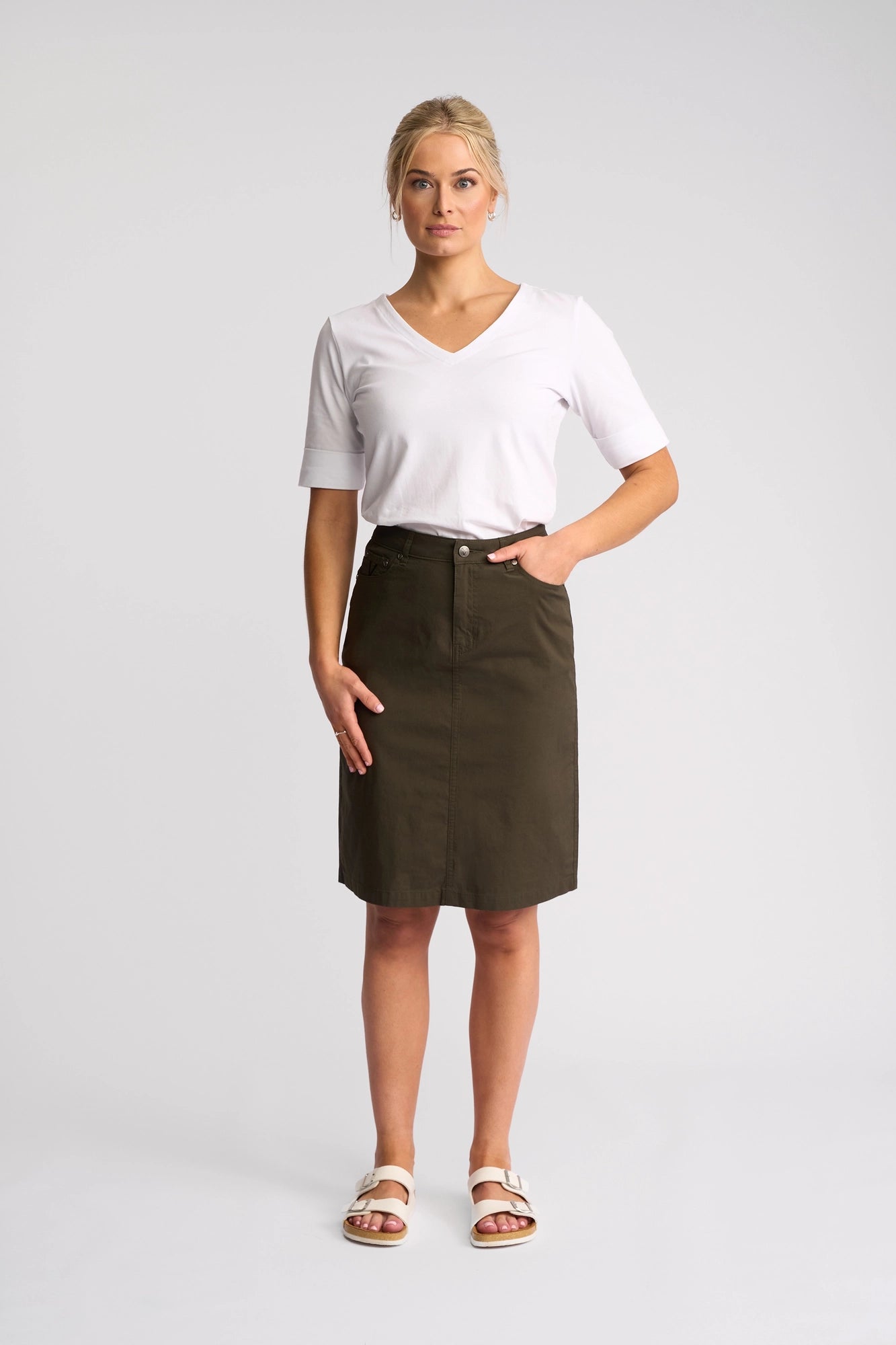 A woman stands facing forward against a plain light background, wearing a white V-neck T-shirt, the Vassalli Poppy Poplin Skirt in Khaki that falls to her knees, and white sandals.