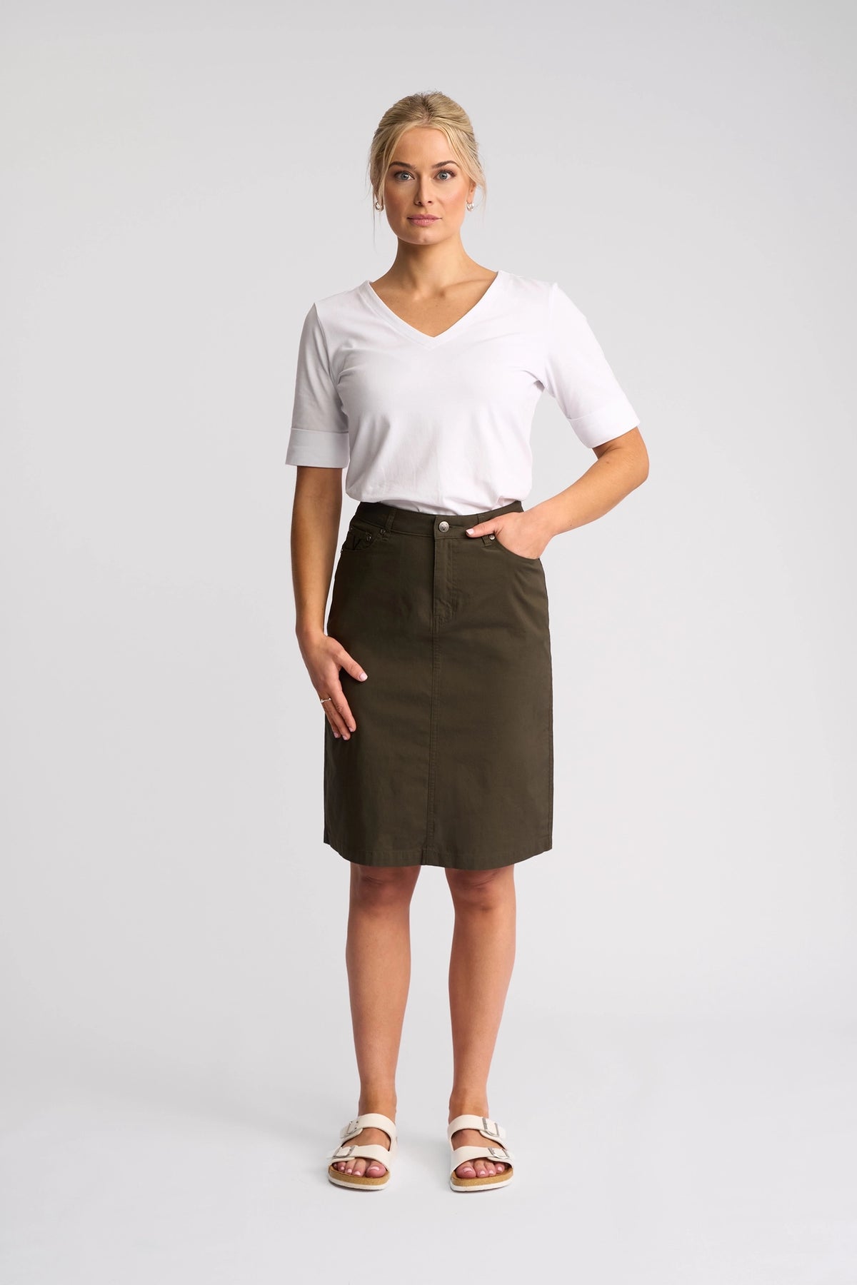 A woman stands facing forward against a plain light background, wearing a white V-neck T-shirt, the Vassalli Poppy Poplin Skirt in Khaki that falls to her knees, and white sandals.