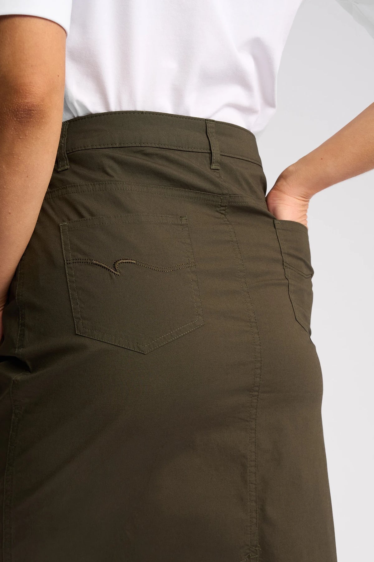 A person wearing the Vassalli Poppy Poplin Skirt in khaki, featuring a back pocket, paired with a white shirt and shown from the waist down with one hand in the pocket.