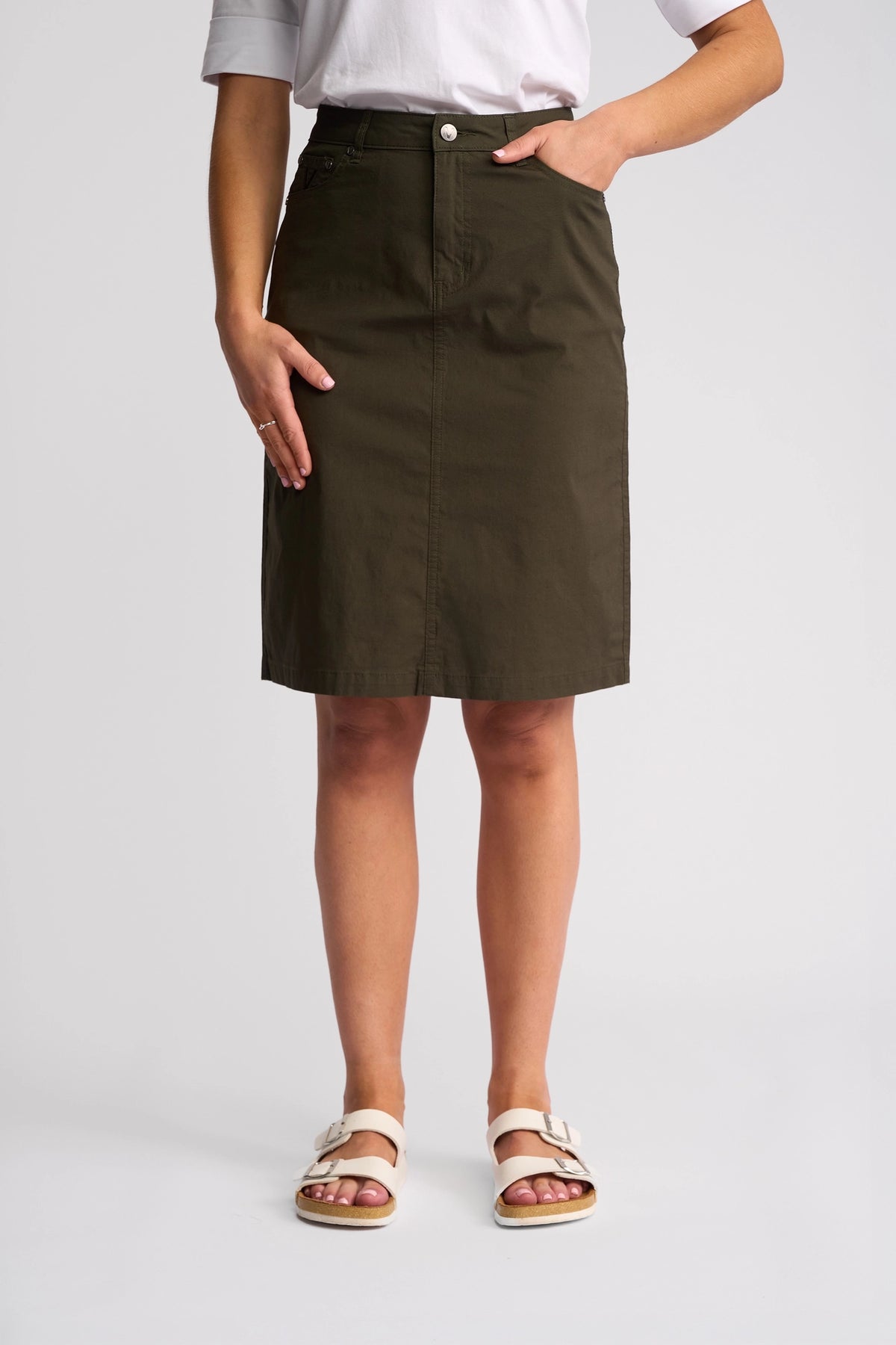 A person stands with one hand in the pocket of the Vassalli Poppy Poplin Skirt in Khaki, paired with a white shirt and white sandals, set against a plain light background.