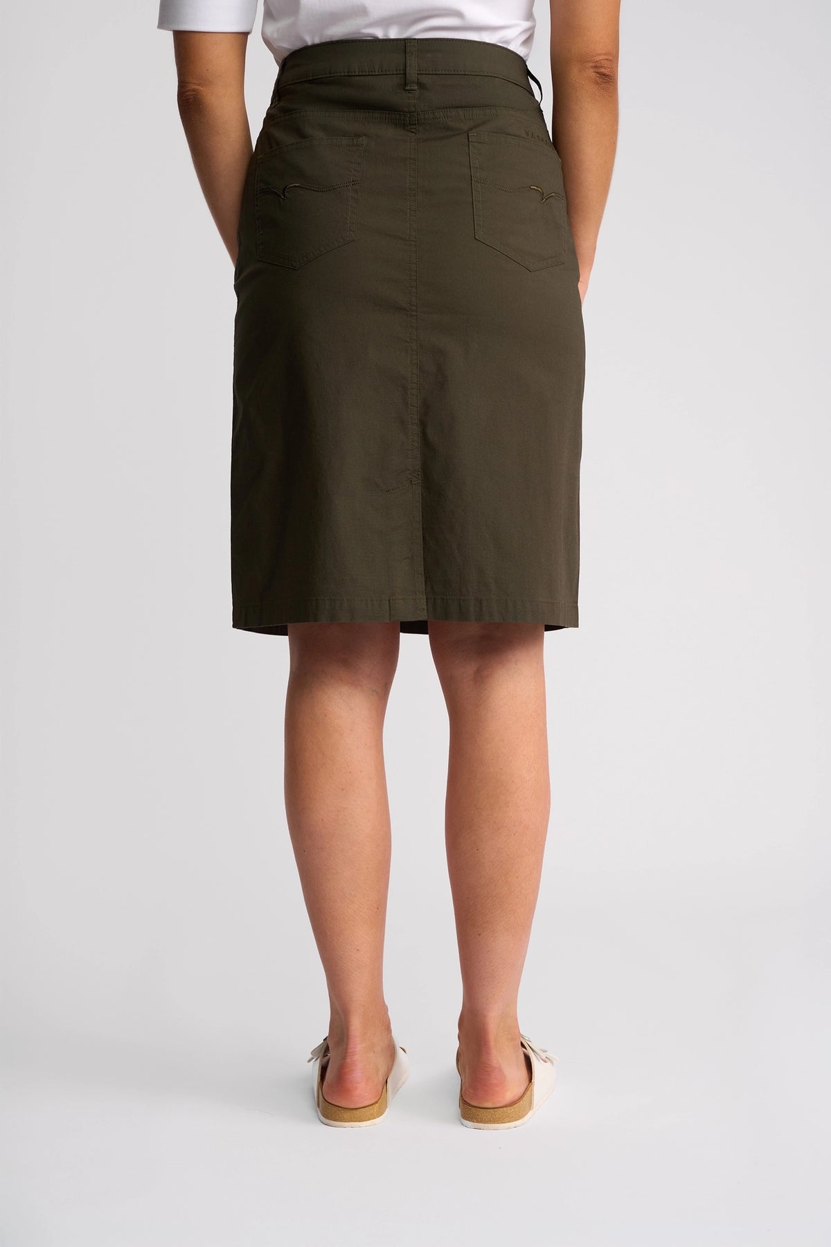 A model is seen from the back against a plain light background, wearing the Vassalli Poppy Poplin Skirt in khaki, paired with a white short-sleeve top and beige slip-on shoes.
