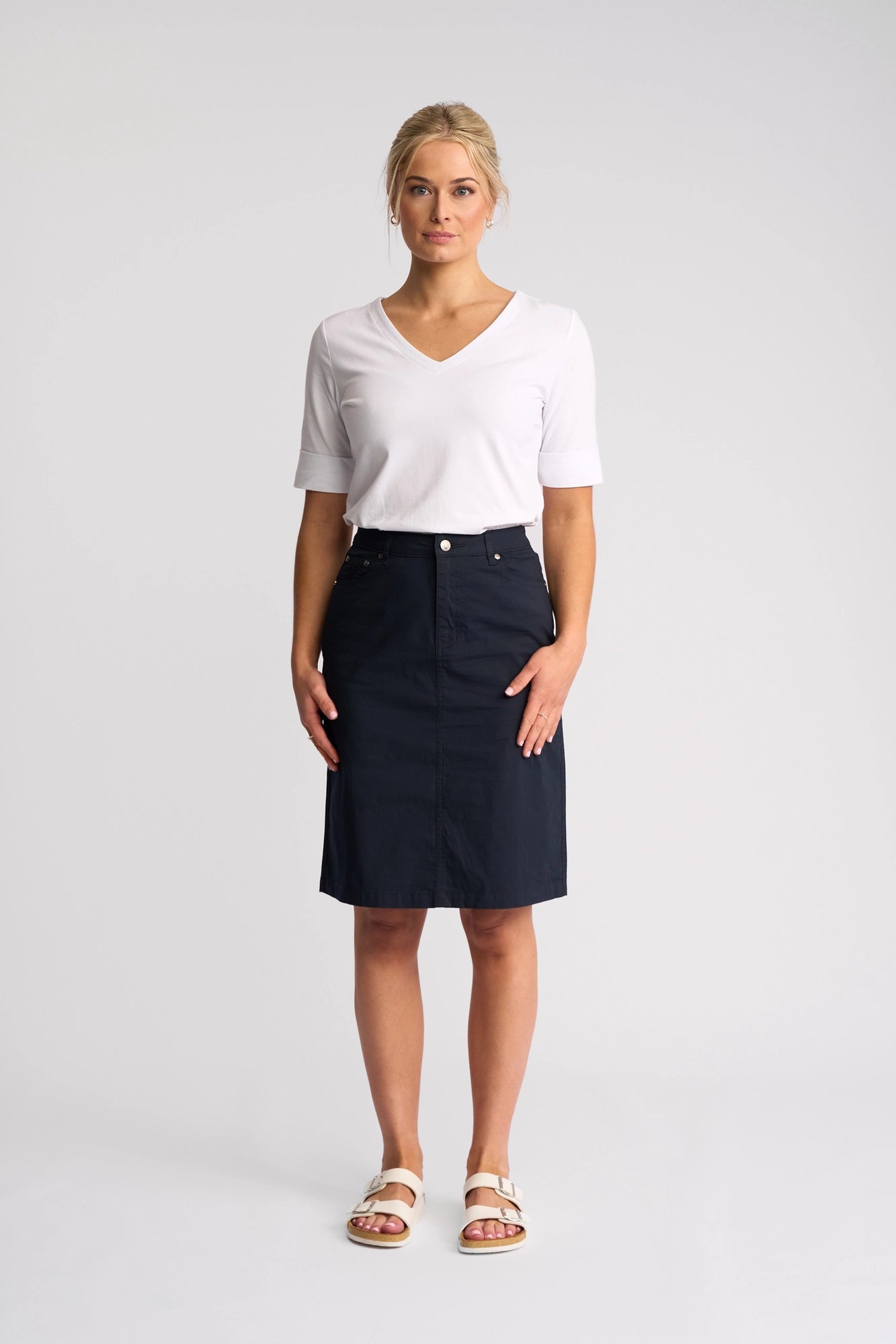 A woman stands against a plain background, wearing a white short-sleeve V-neck shirt, the Vassalli Poppy Poplin Skirt in Navy that falls to the knee, and white sandals, looking straight at the camera with a neutral expression.
