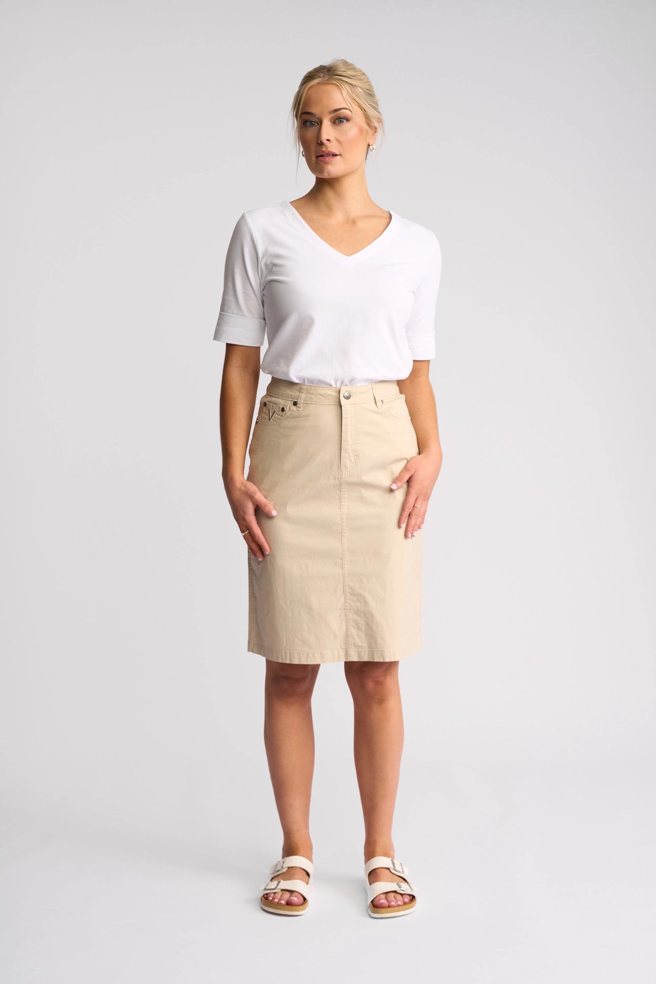 A woman stands against a plain background, wearing a white V-neck short-sleeve top, the Vassalli Poppy Poplin Skirt in Sand crafted from breathable fabric, and white sandals. Her blonde hair is pulled back as she gazes slightly to the side.