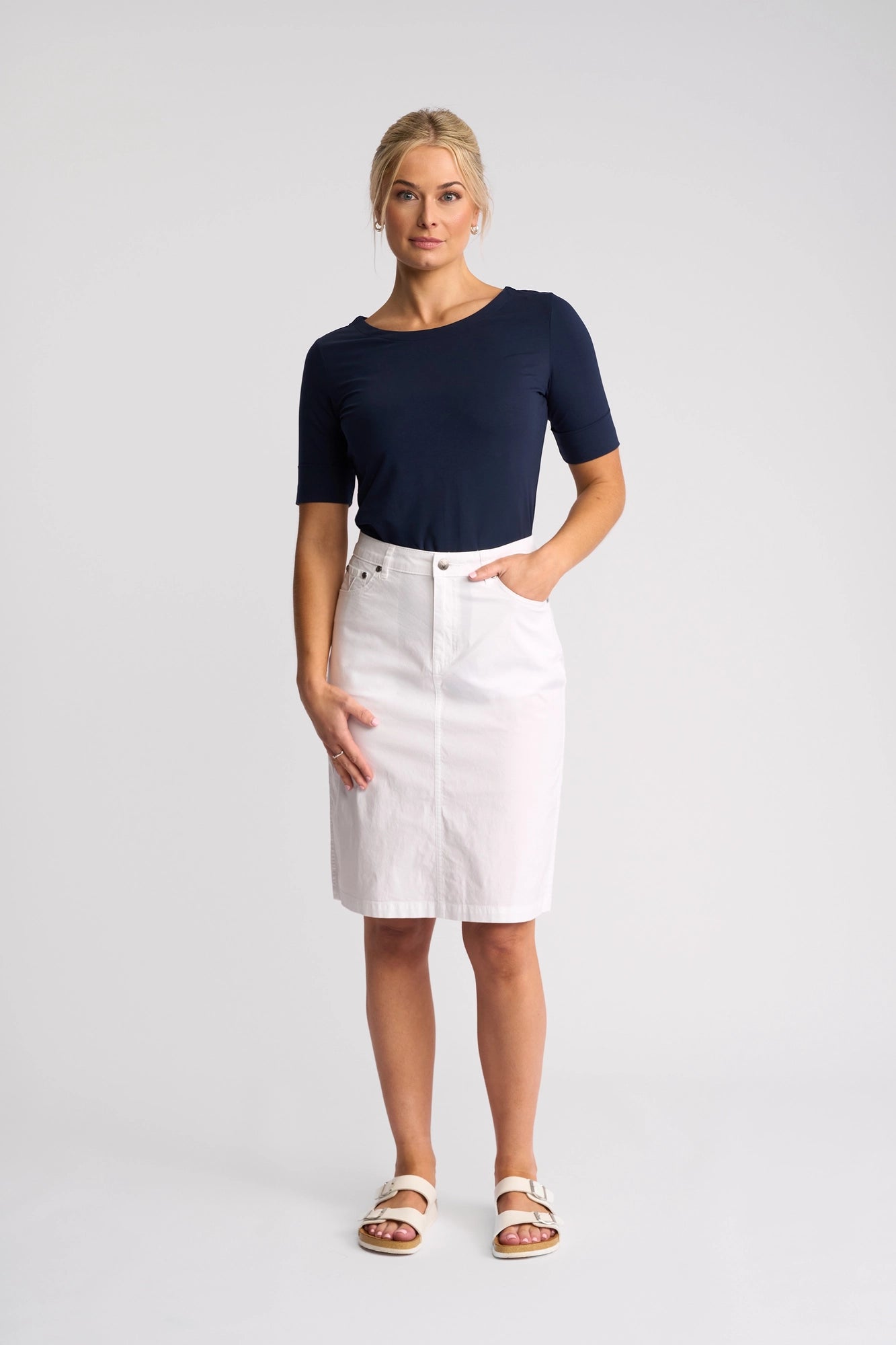 A woman stands against a plain background, wearing a navy blue short-sleeve top, white sandals, and the Vassalli Poppy Poplin Skirt in White—a crisp pencil skirt that’s perfect for summer. She poses with one hand in her pocket and a neutral expression.