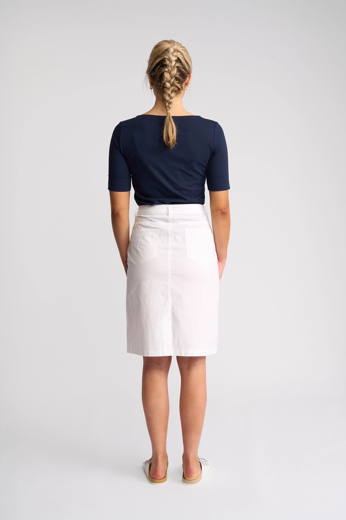 A woman with a blonde braid stands facing away, wearing a navy short-sleeve top, the Vassalli Poppy Poplin Skirt in White featuring a back pocket, and light-colored slip-on shoes, against a plain white background.