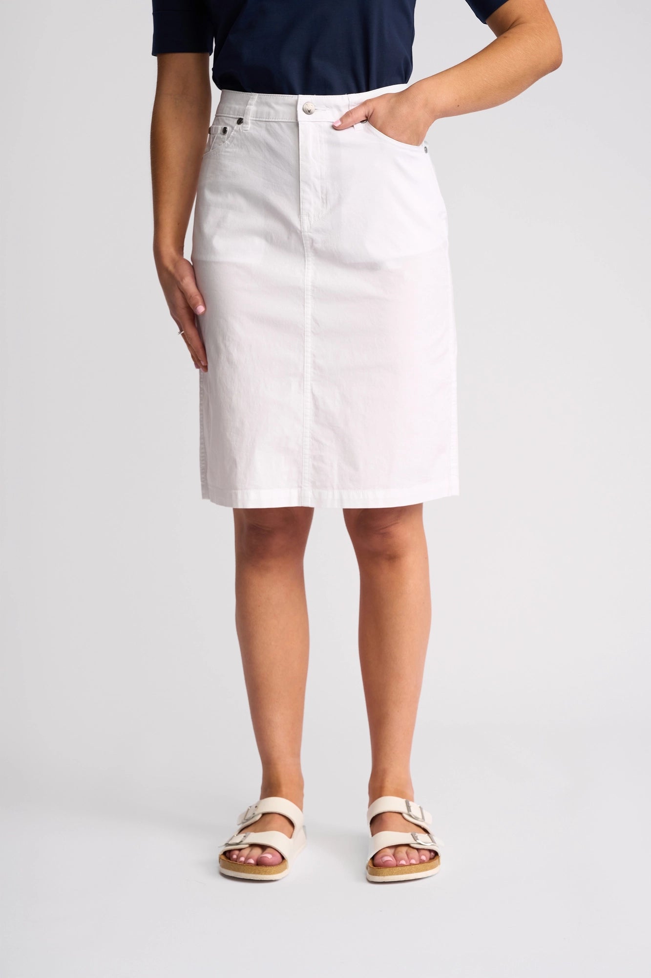 A person stands against a plain background wearing a navy top, white Vassalli Poppy Poplin Skirt featuring front pockets, and white sandals, with one hand in a pocket.
