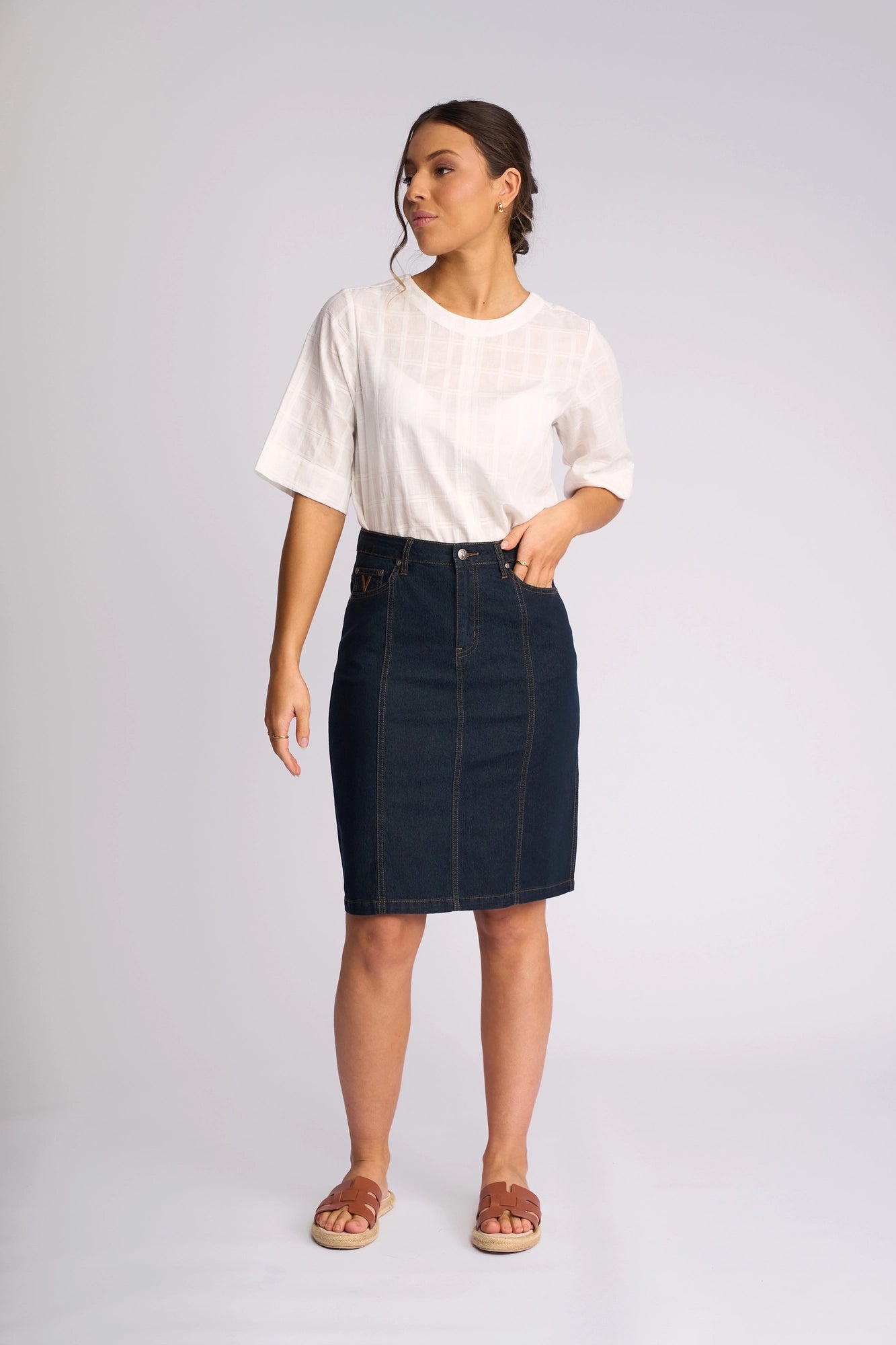 A woman stands against a light background wearing a white short-sleeve shirt, Vassalli’s Poppy Skirt in Indigo Denim, and sandals. She looks to the side with one arm relaxed and the other slightly bent.