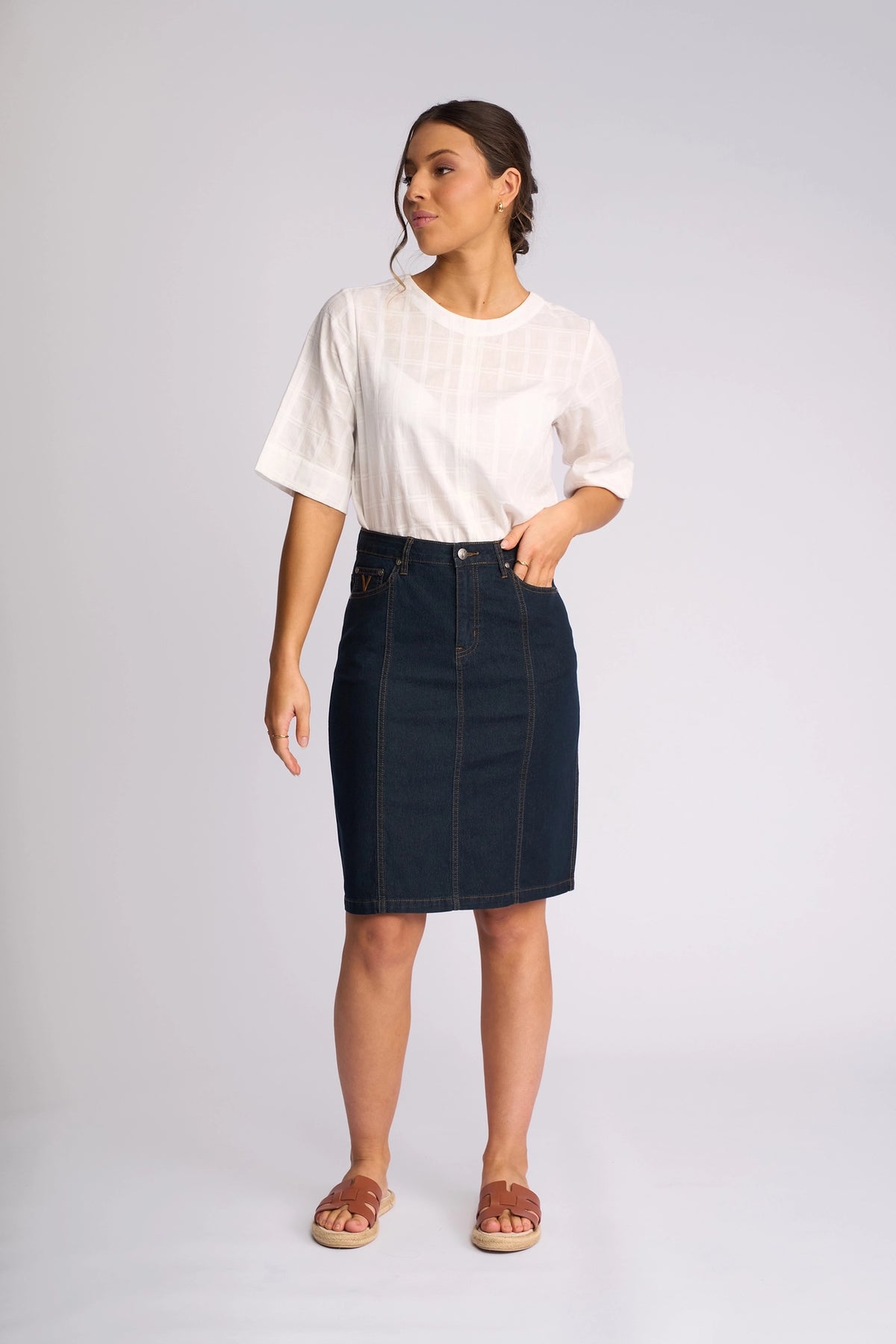 A woman stands against a light background wearing a white short-sleeve shirt, Vassalli’s Poppy Skirt in Indigo Denim, and sandals. She looks to the side with one arm relaxed and the other slightly bent.