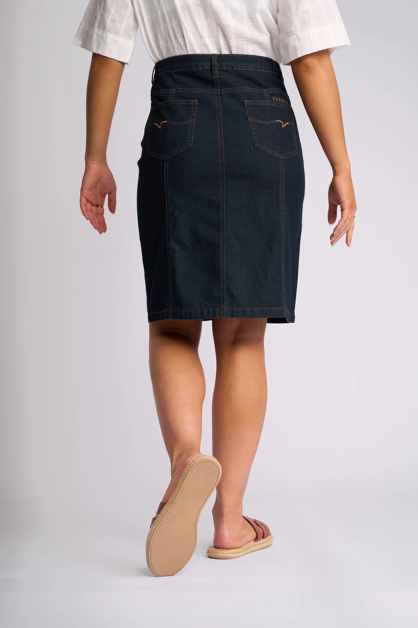 A person wearing a white shirt, the Vassalli Poppy Skirt in Indigo Denim that falls to the knees, and sandals is standing with their back to the camera against a plain background.