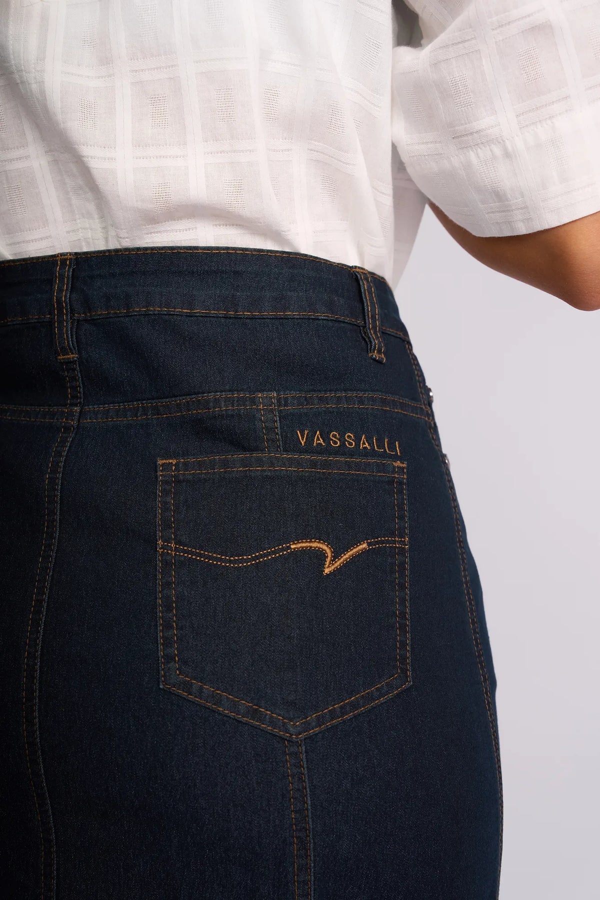 A person wearing the Vassalli Poppy Skirt in Indigo Denim, featuring distinctive Vassalli embroidery above the back pocket, paired with a white textured shirt. The image highlights the skirt’s pocket and branding, capturing a timeless look reminiscent of classic indigo denim styles.