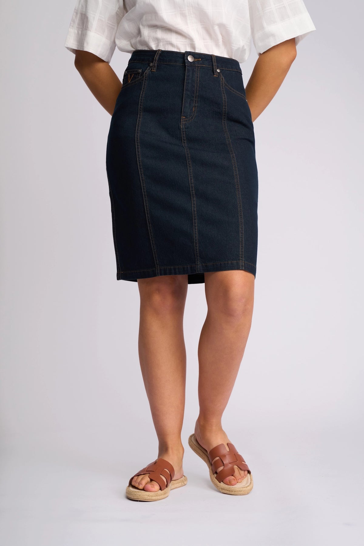 A person wearing a white short-sleeve shirt, the Vassalli Poppy Skirt in indigo denim, and brown sandals stands against a plain light background.